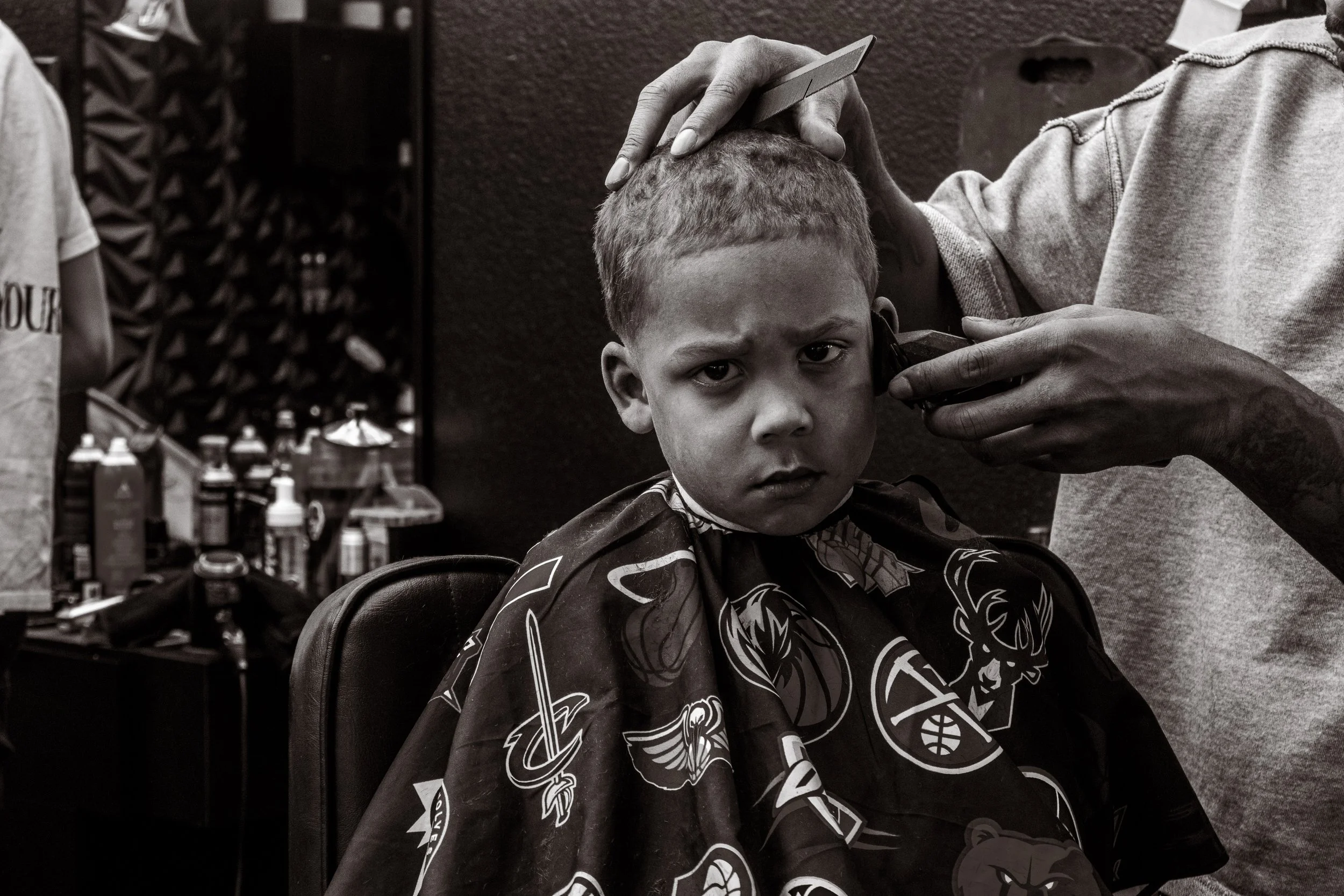 A young boy getting a haircut at a barber shop, looking serious and slightly annoyed.