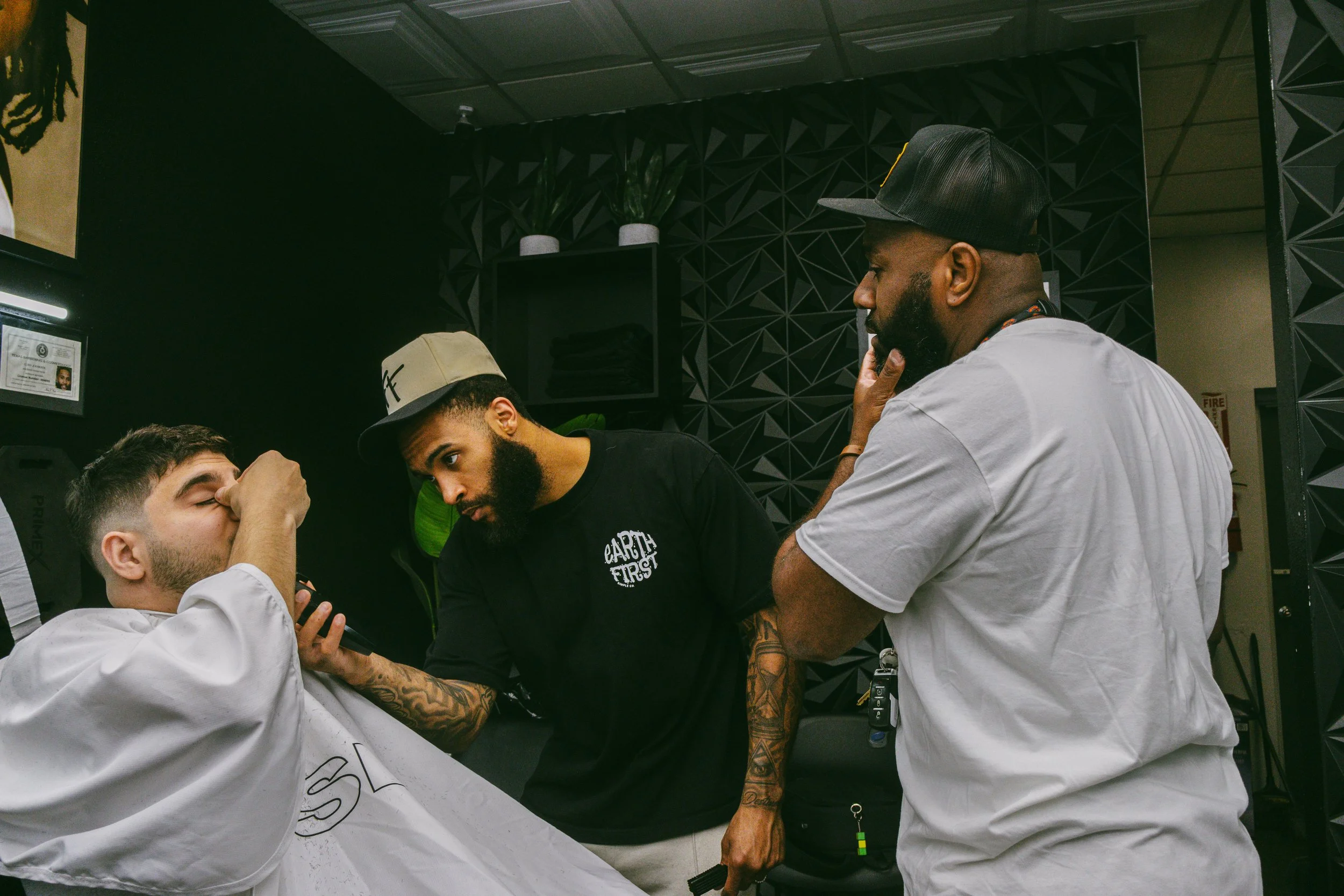 A barber trims a customer's eyebrow while two other barbers observe.