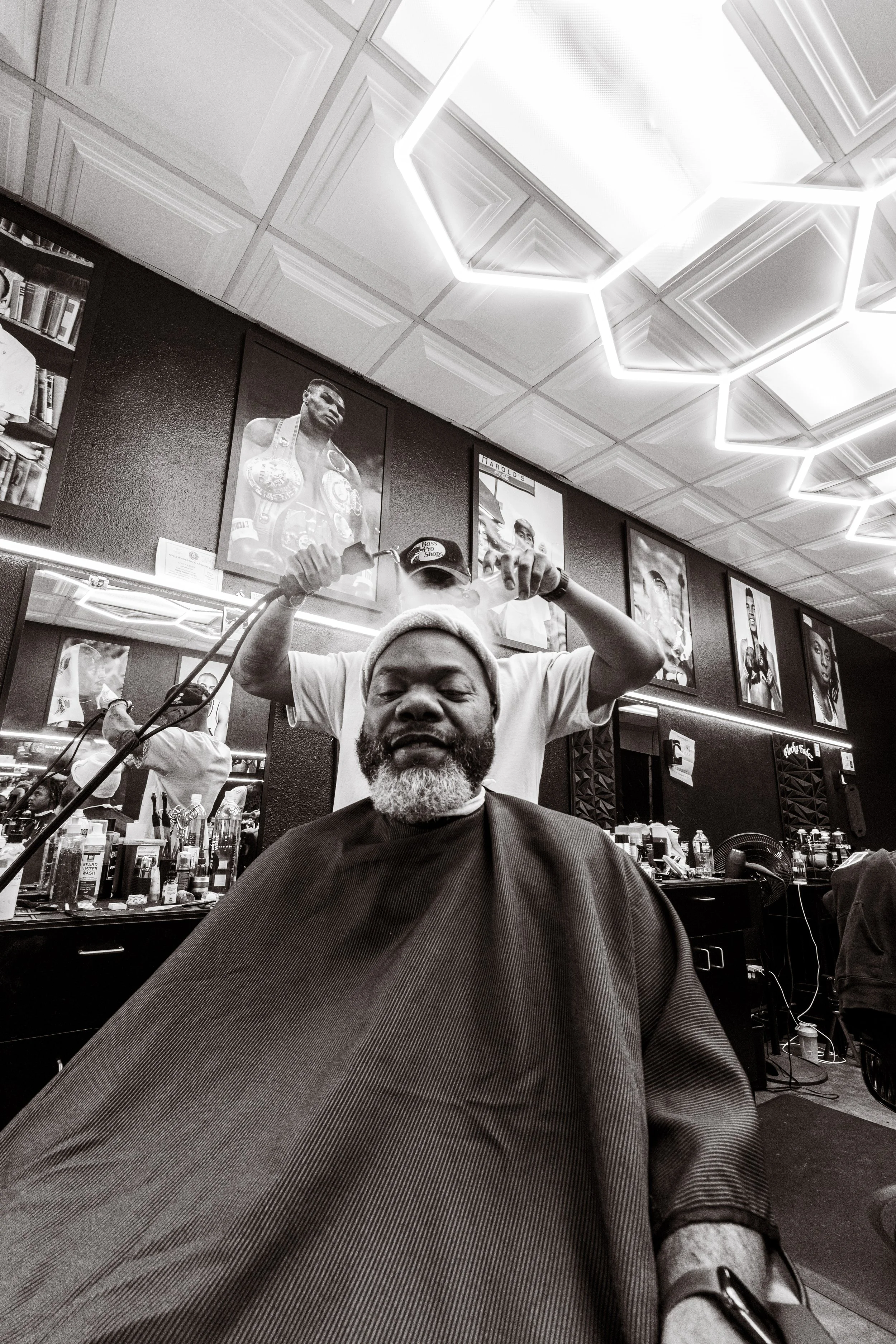 A man with a beard and a headband sitting in a barbershop chair, getting a haircut. The barbershop has framed boxing photographs on the wall and modern geometric lighting on the ceiling.