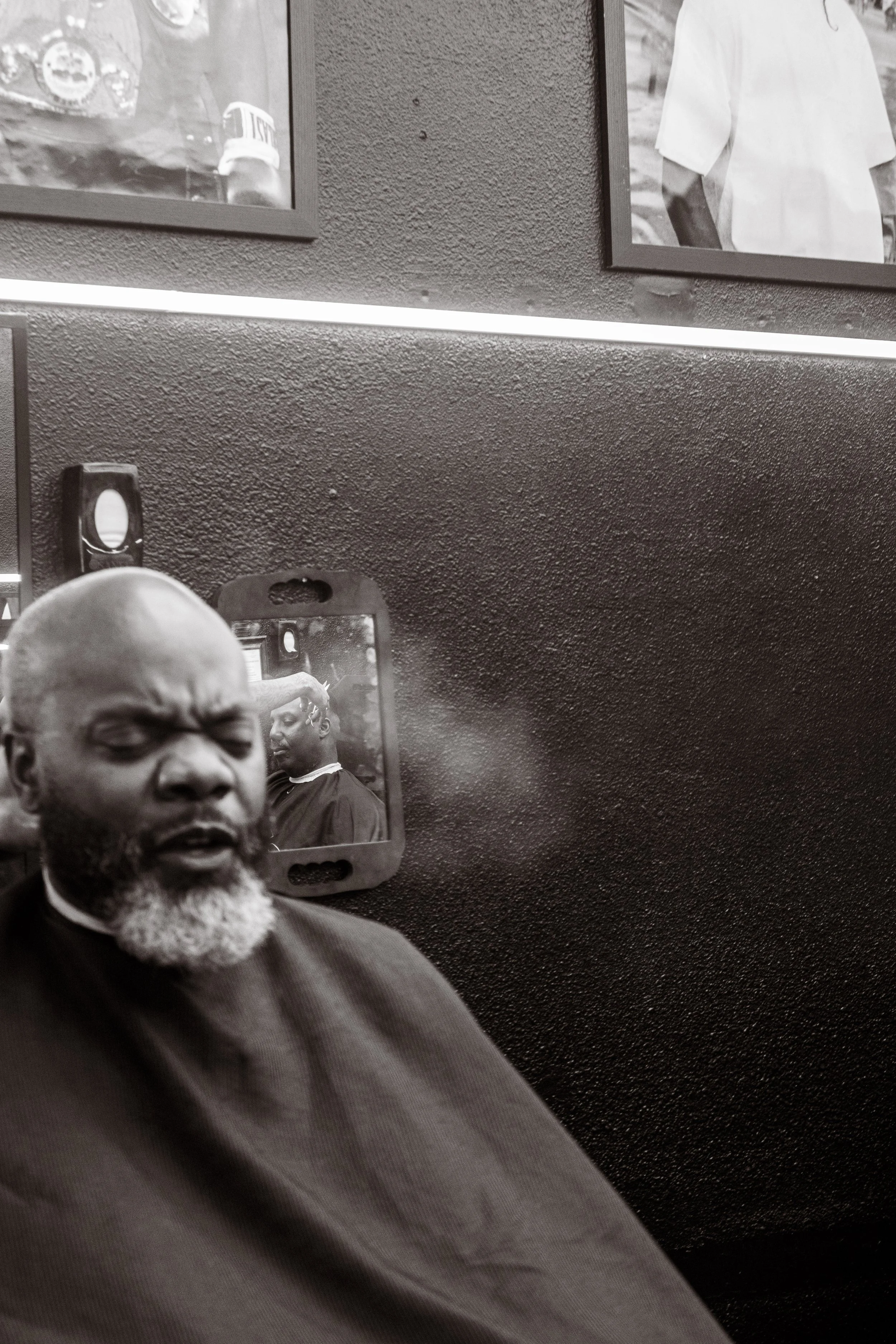A man with a beard sitting in a barbershop, reflected in a mirror behind him. The background features framed photos on a textured black wall.
