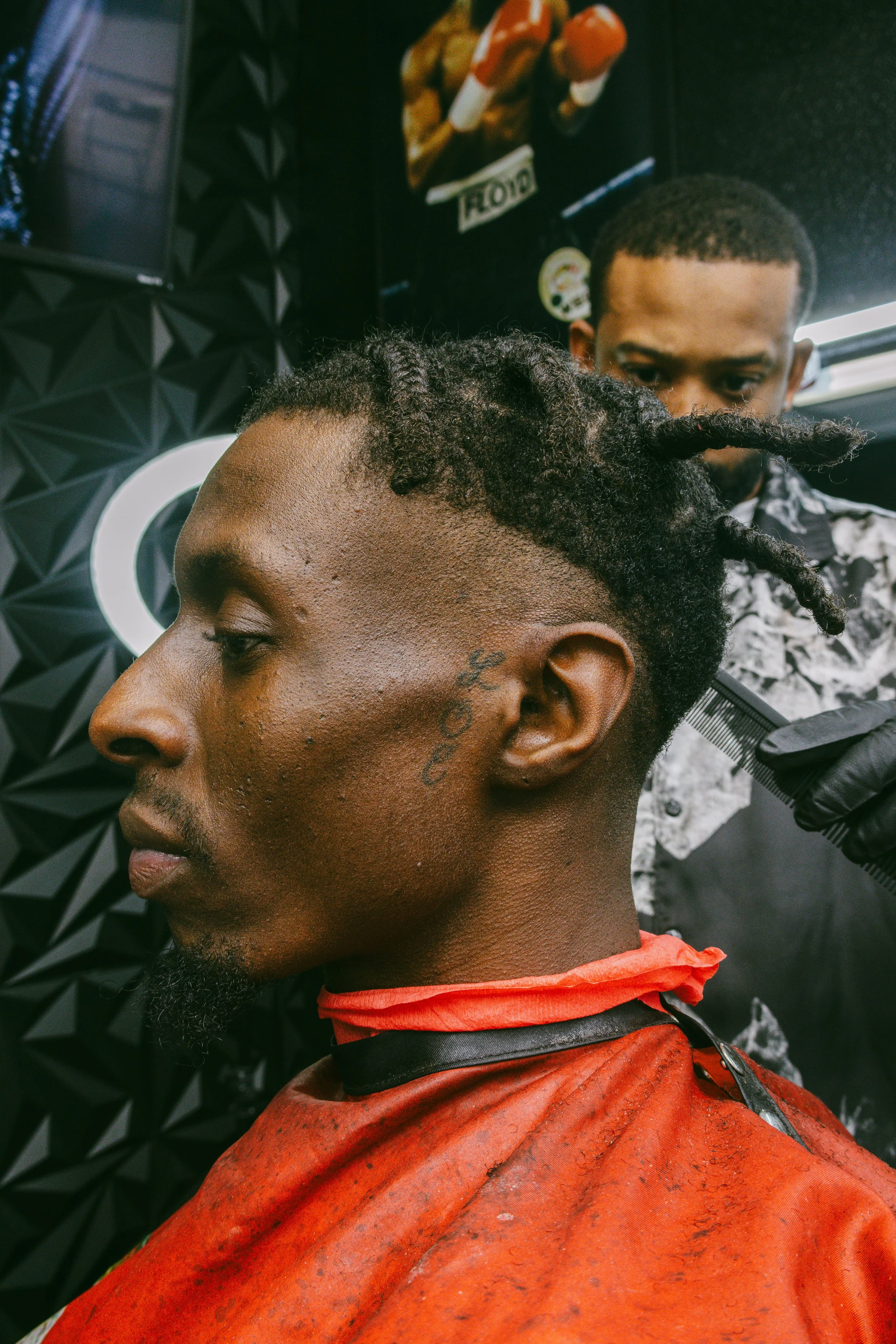 A man getting a haircut with the word 'COOL' tattooed on his face, wearing a red barber cape, and seated in a barbershop with a stylist behind him.
