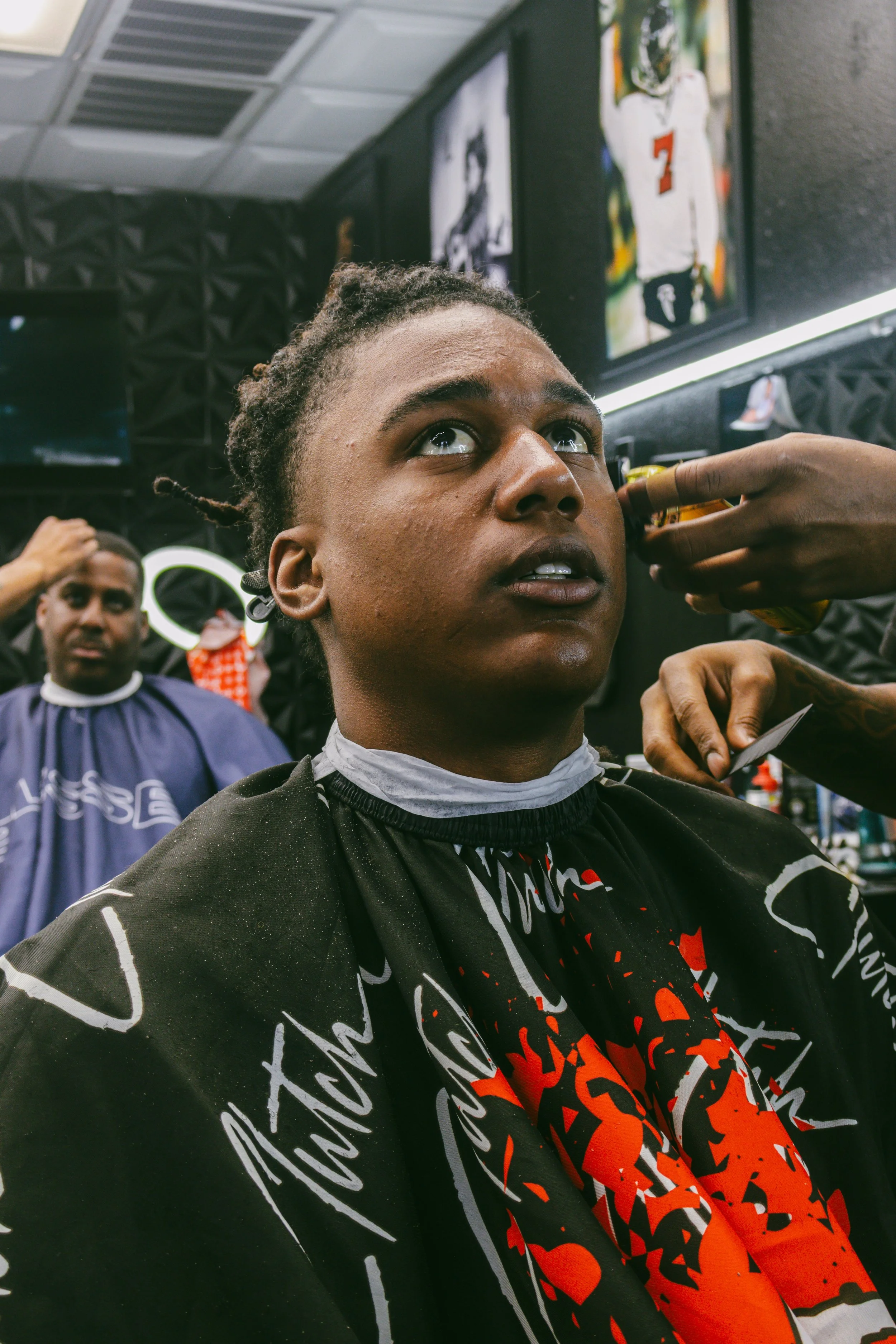 A young man getting a haircut at a barbershop, with another person visible in the background.
