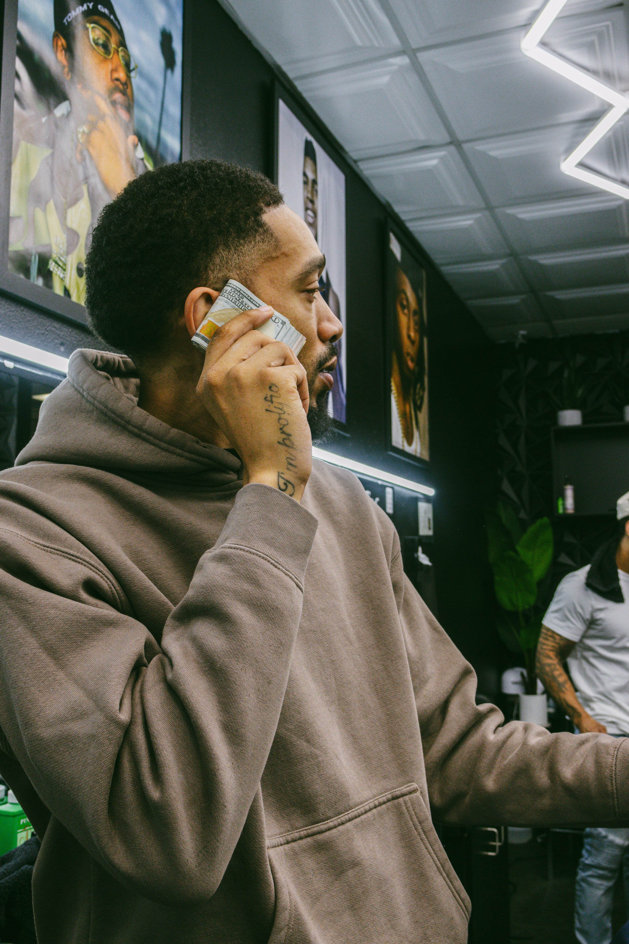 A man with tattoos on his hand talking on a cell phone in a tattoo studio, with colorful portraits on the wall behind him.