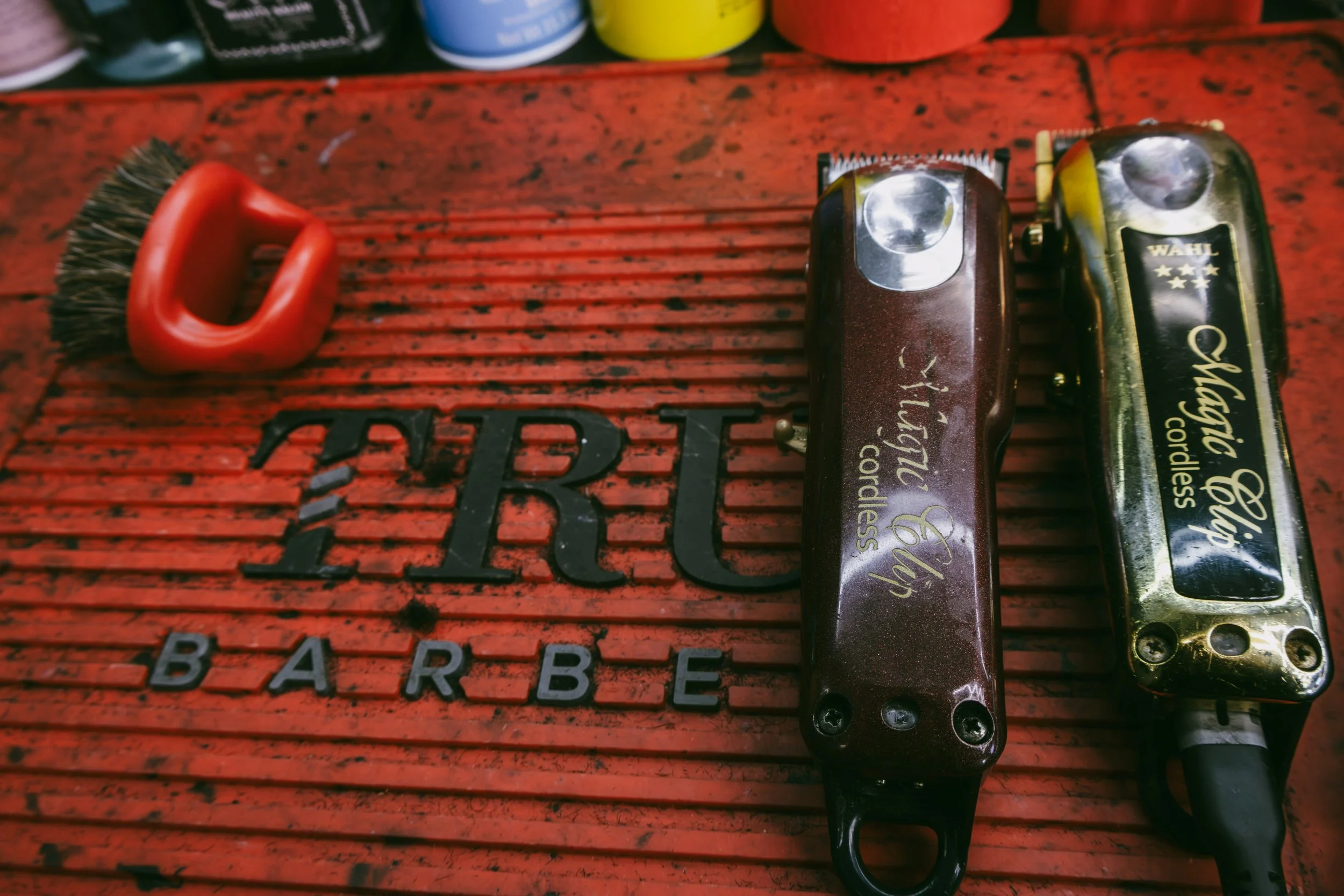 A red barber shop mat with the words "BARBER" printed on it in black and white, a wire brush with a red handle, and two electric hair clippers, one brown and one gold, placed on the mat.