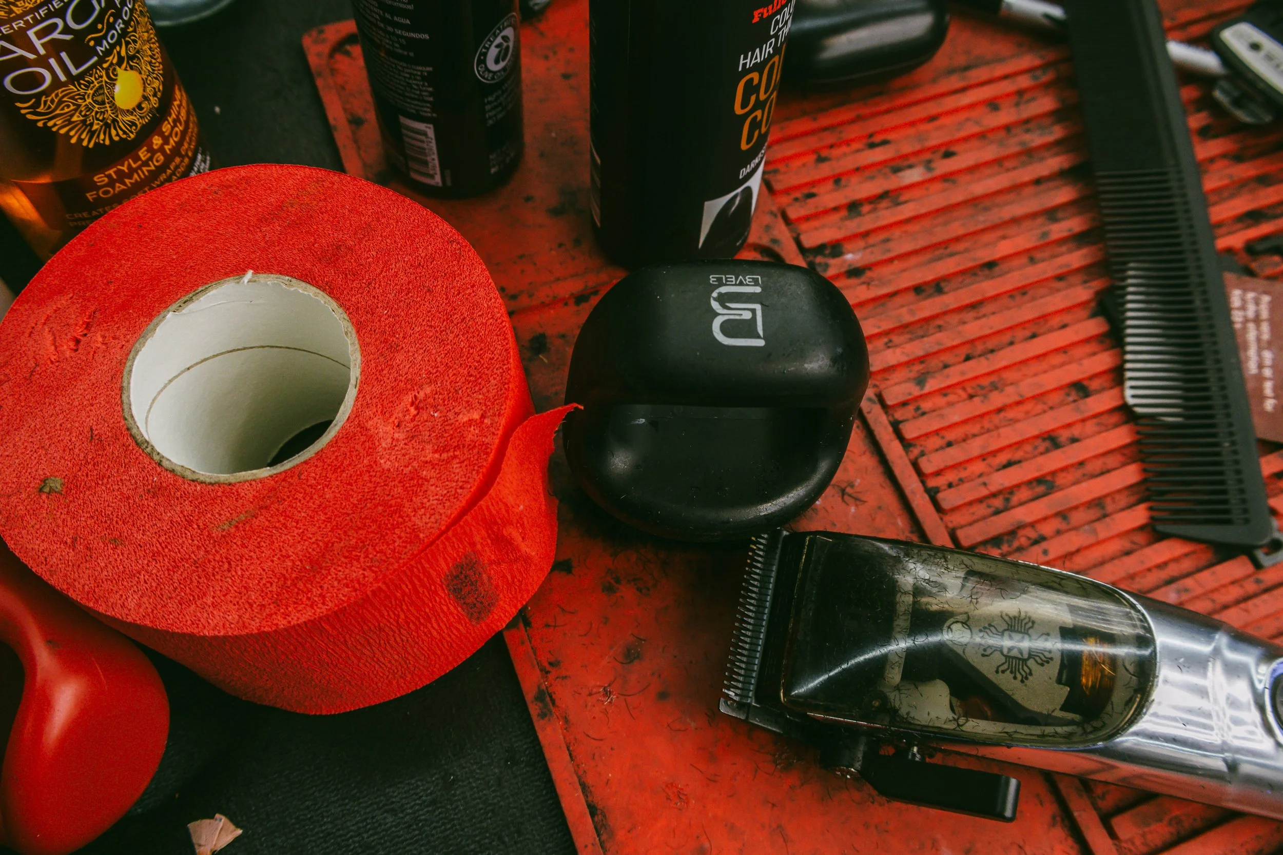 A red roll of cloth tape, a black wireless earbud case, a pair of electric hair clippers, a black comb, and various grooming products on a red work surface.