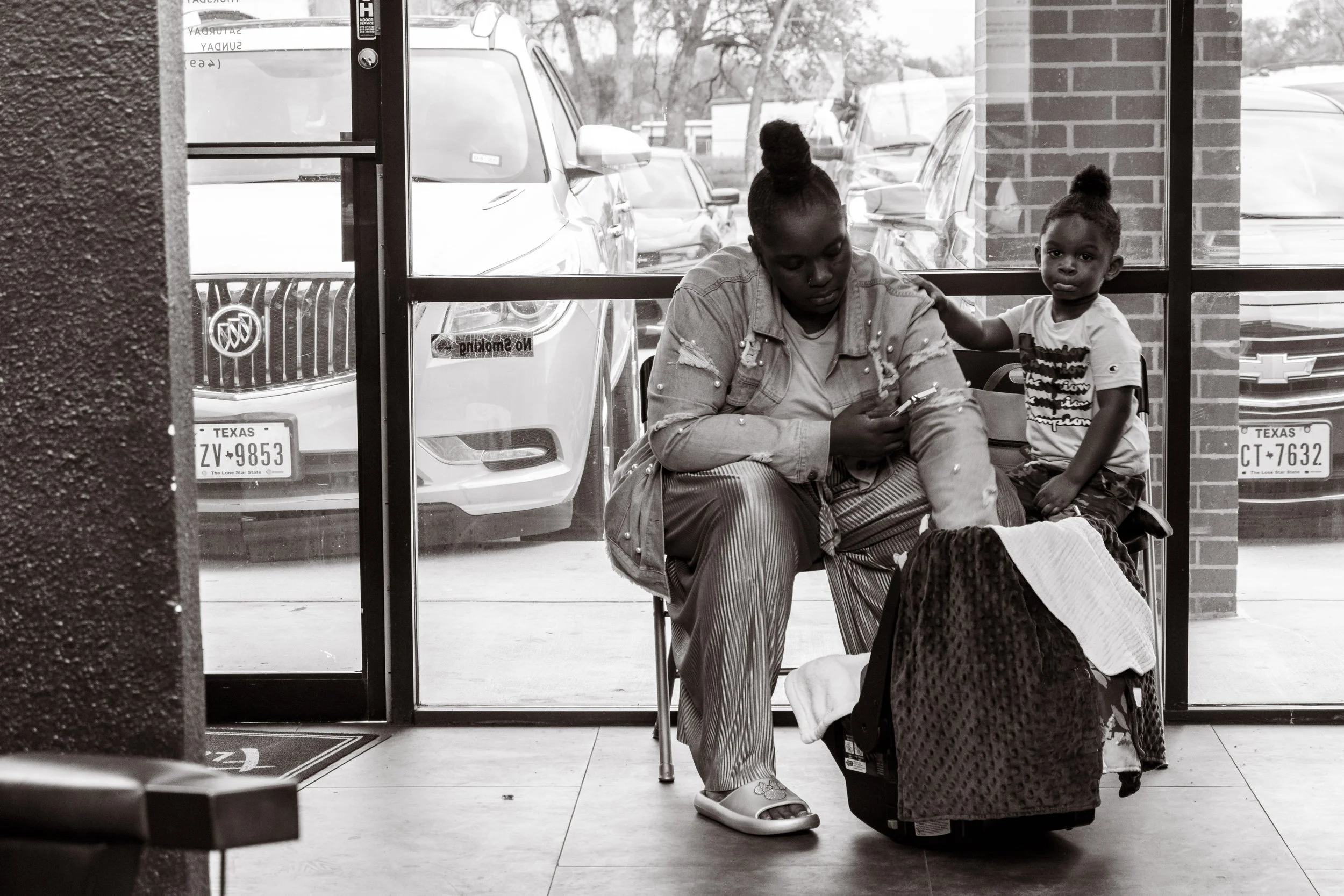A woman and a young girl sitting inside a building near a glass door, with cars visible outside. The woman is looking at her phone, while the girl looks directly at the camera, touching the woman's arm, with a bag and blankets on her lap.