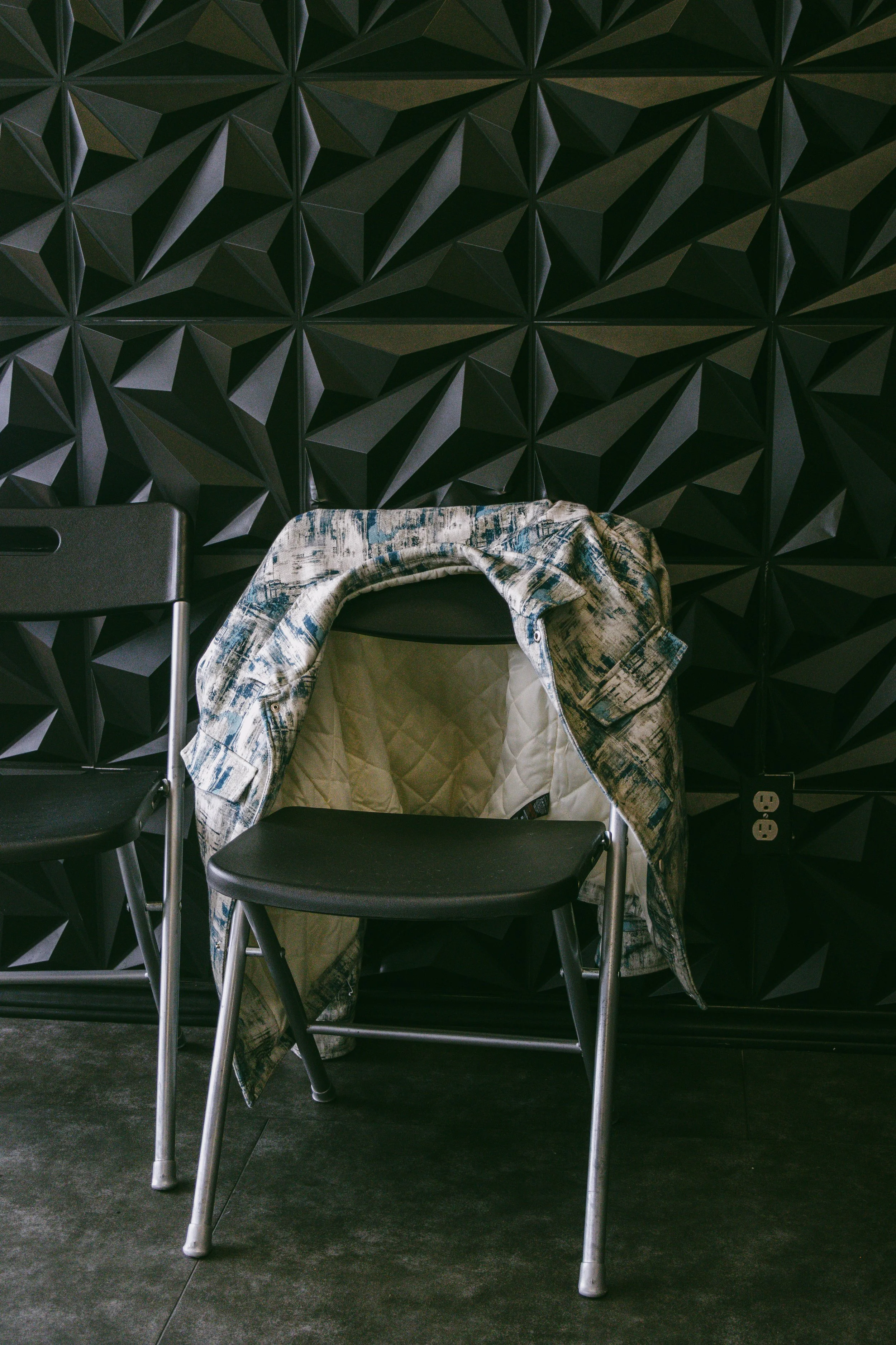 Black chair with a plaid jacket draped over it, against a textured black wall with geometric patterns, and electrical outlet nearby.