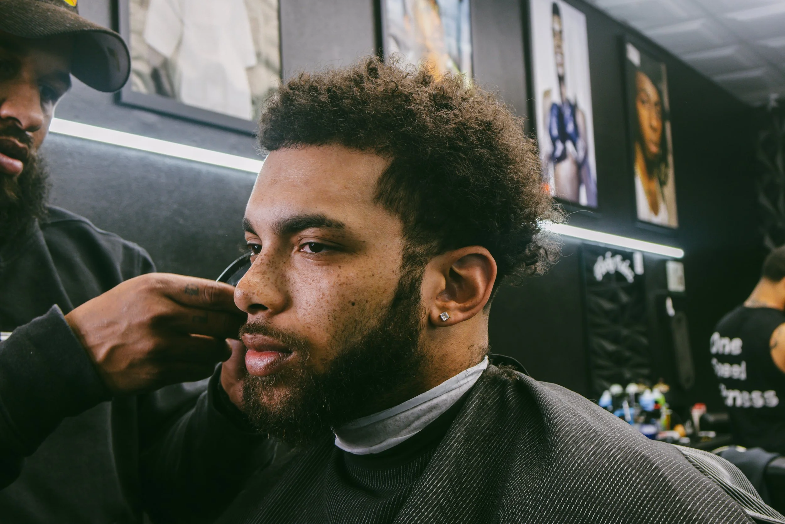 Man with curly hair and beard getting a haircut at a barbershop while barbershop mirrors and artwork are visible in the background.