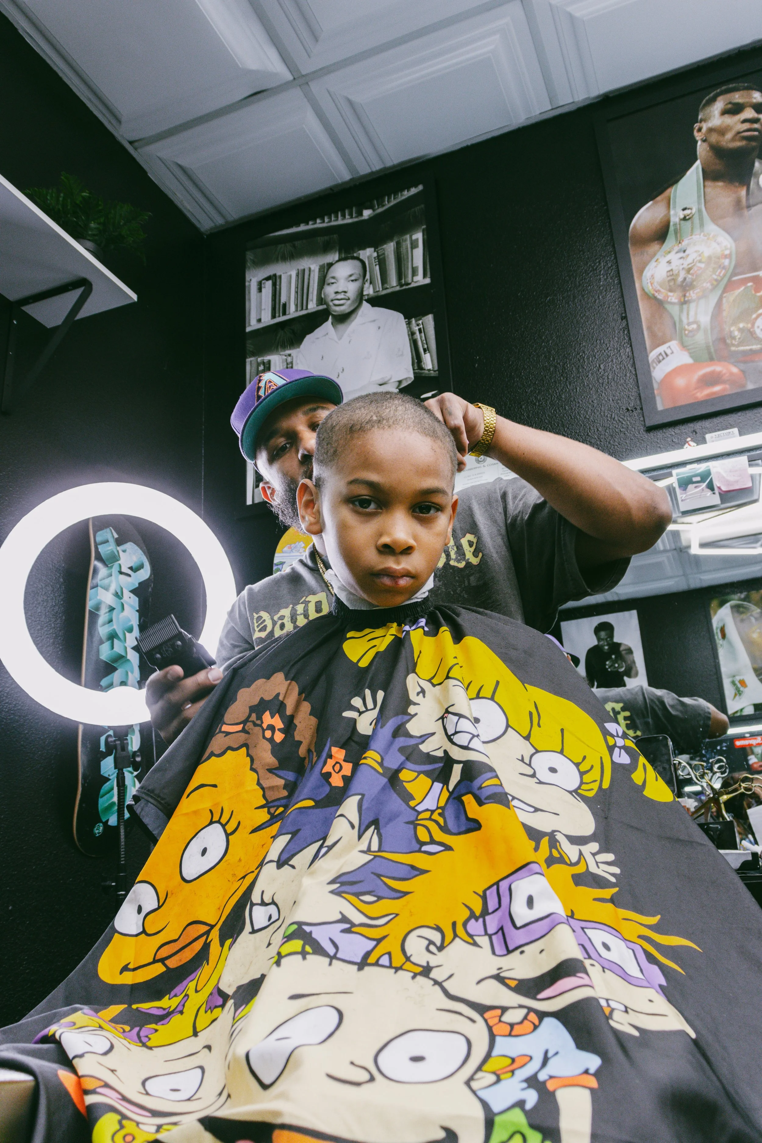A young boy getting a haircut in a barbershop, sitting under a cape decorated with cartoon characters. A barber is behind him, trimming his hair with a clipper. The background shows black walls with framed photos and a bright ring light.
