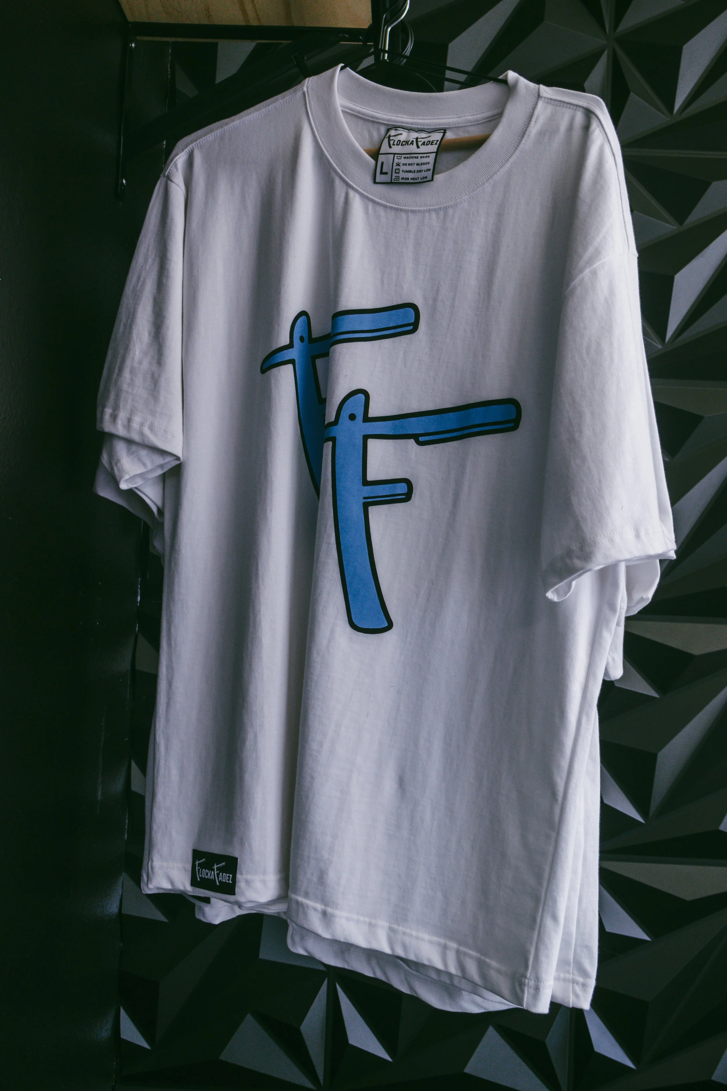 A white T-shirt hanging on a black hanger. The shirt has a graphic design of two blue spray paint cans with black outlines. The background features a geometric-patterned wall.
