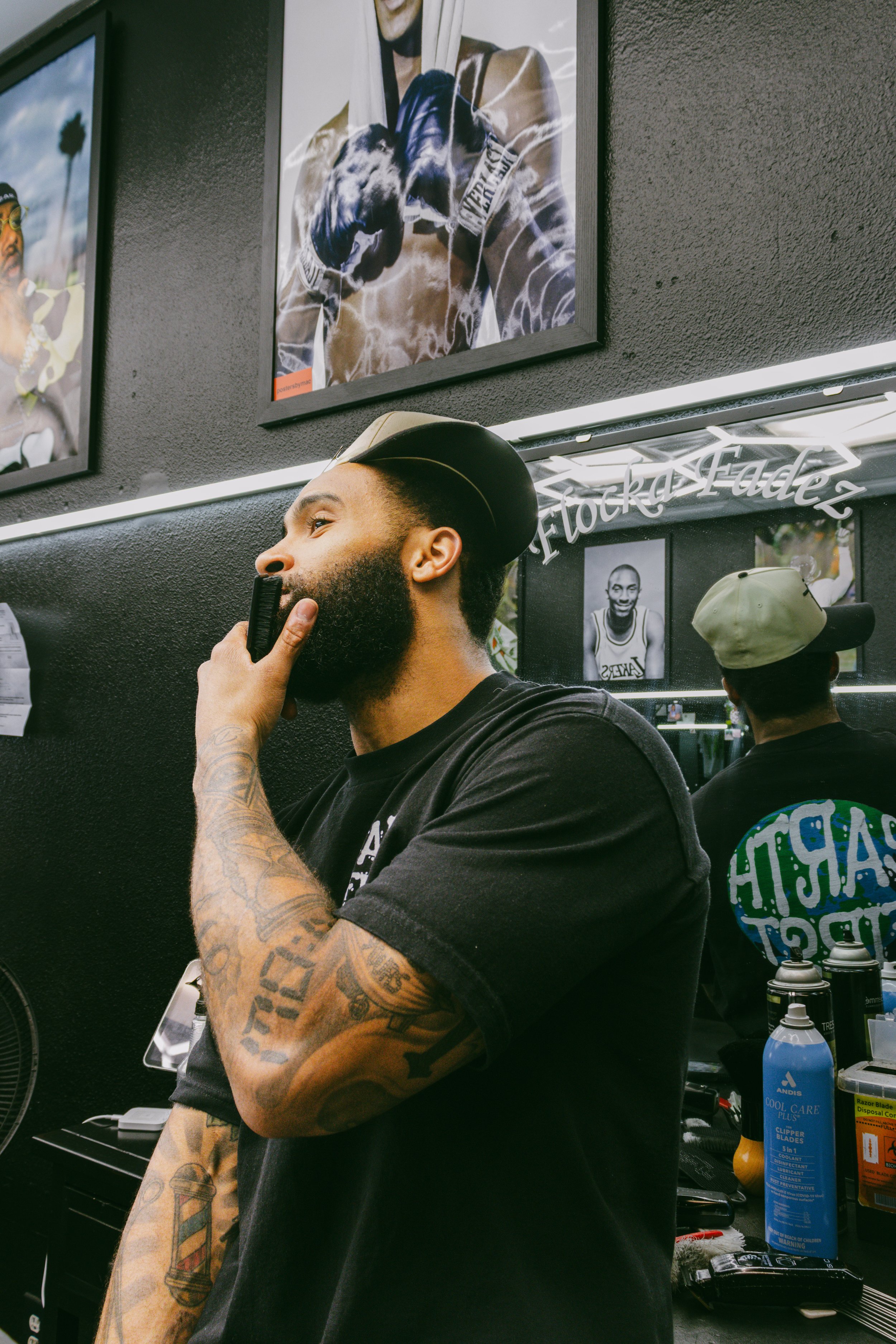 A bearded man with tattoos on his arm is sitting in a barbershop, holding a straight razor to his face. He is wearing a black t-shirt and a black cap. The shop has black walls decorated with framed photos and artwork, including a portrait of a person
