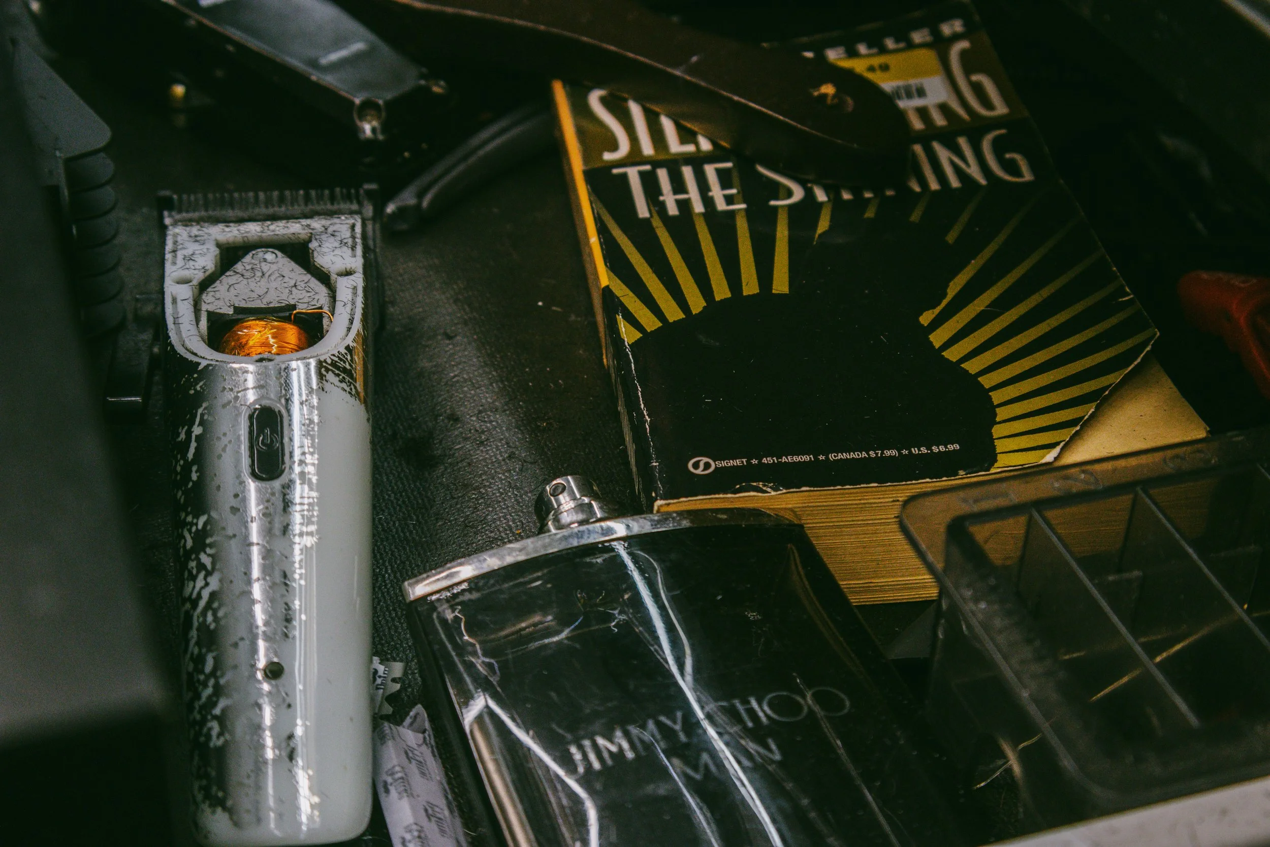 Close-up view of a disassembled electric razor, a worn Jimmy Choo box, and a black container with a screw lid on a textured surface.