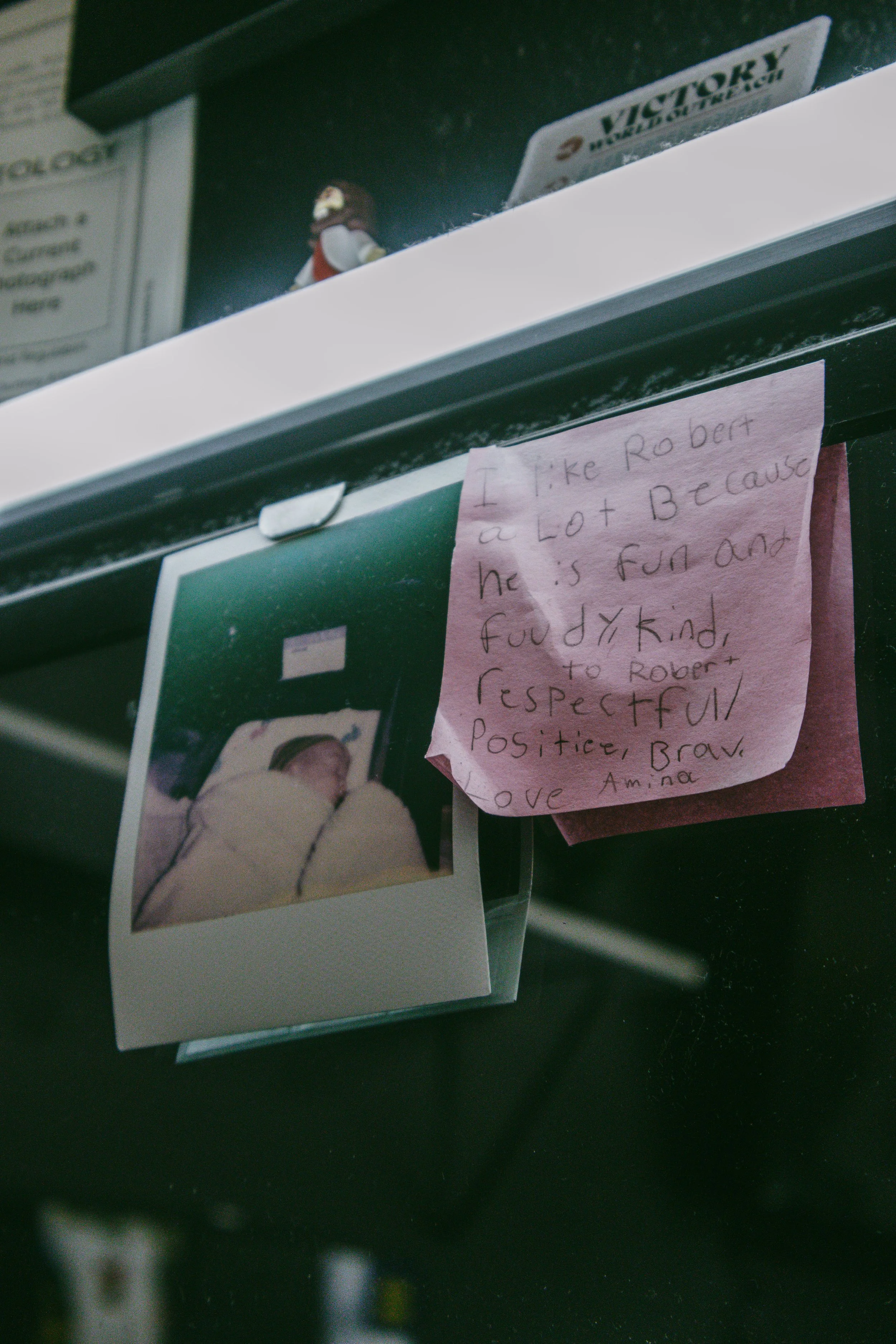 A handwritten note on pink paper expressing admiration for Robert, praising his fun and kind personality, and love from Amina. A photograph of a person in a hospital bed is attached below the note.