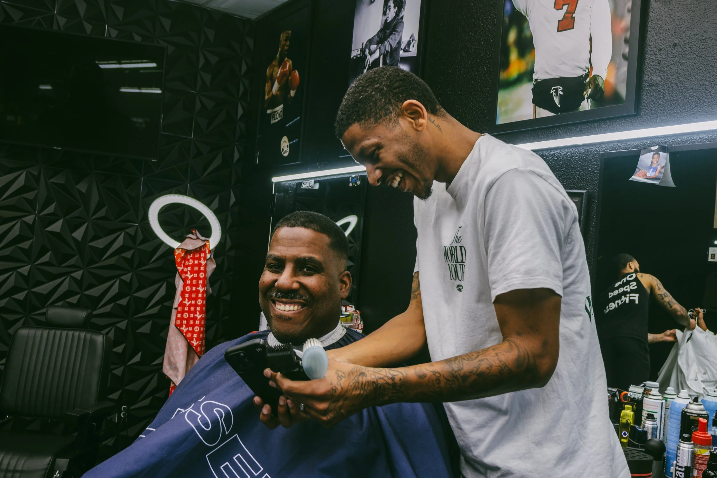 A young man getting a haircut in a barbershop, smiling and looking at his phone while a barber is styling his hair, both smiling and appearing happy.