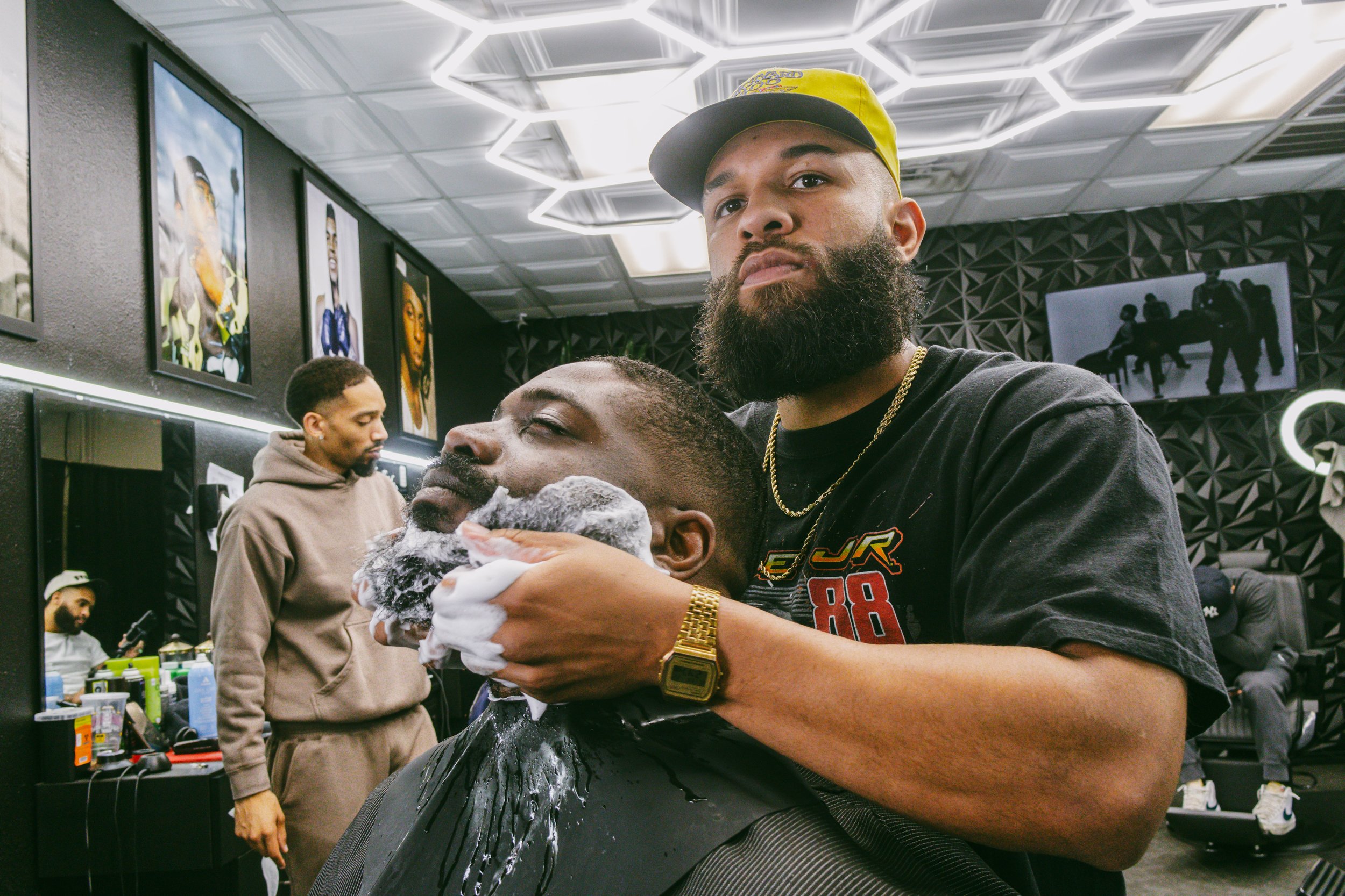 A man with a beard getting a shave at a barber shop, with another man standing in the background and a woman sitting and looking at her phone.