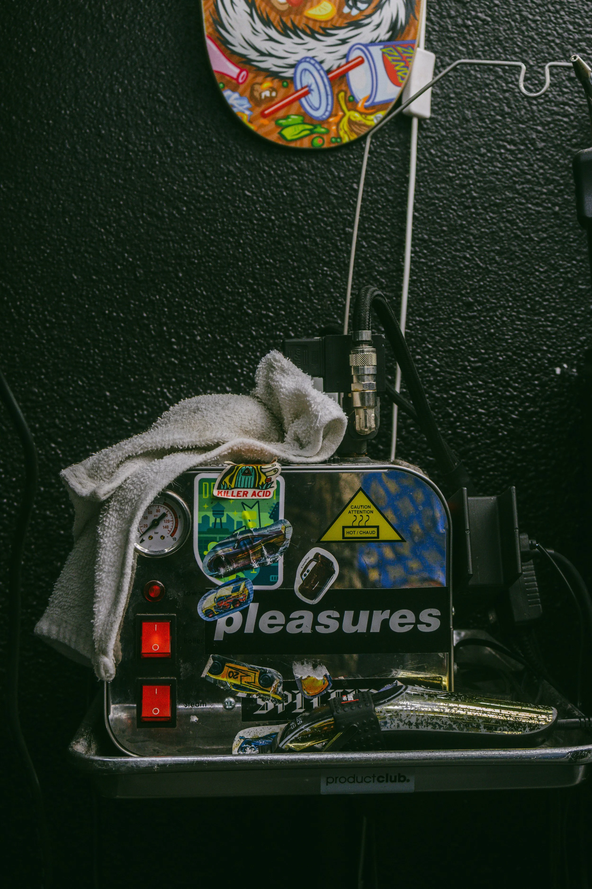 A metallic box with various stickers and switches, a white towel, and a power cord, positioned against a black textured wall. A colorful circular artwork hangs above.