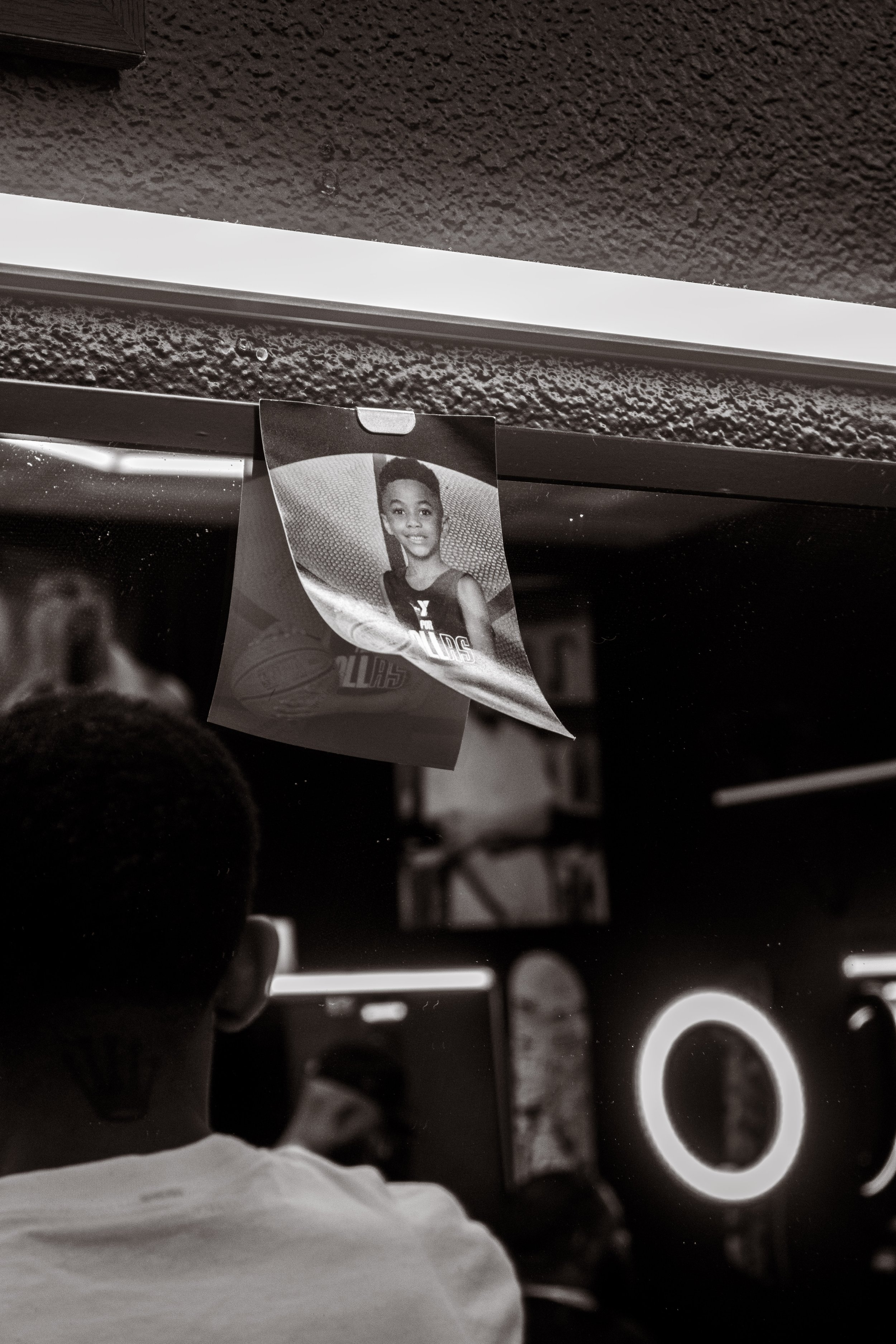 A black and white photo of a teenager in a basketball jersey, reflecting in a mirror, with part of a photograph of the same teen hanging above.