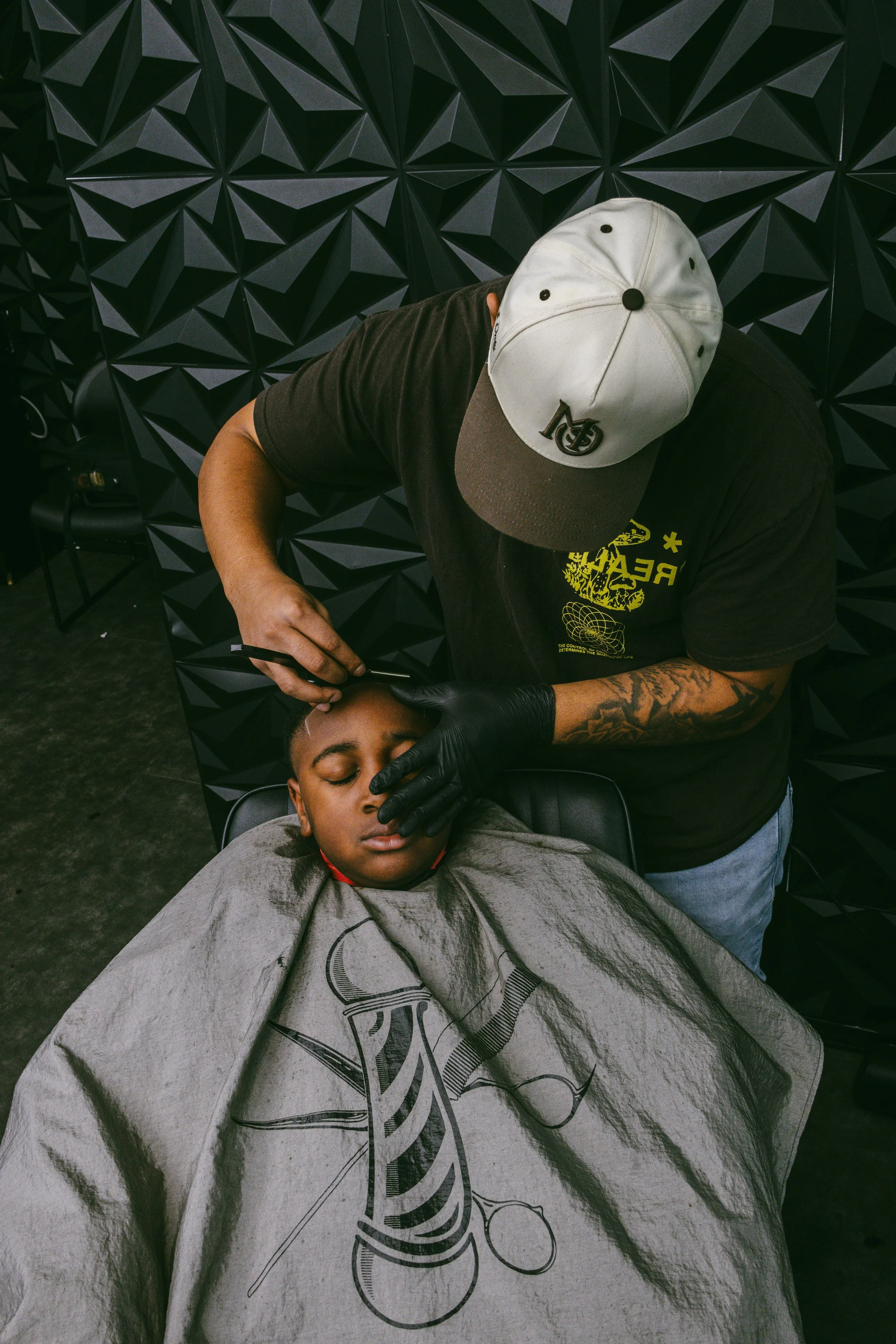 A young boy is getting a haircut in a barbershop, lying back in a chair with a barber trimming his hair. The barber is wearing a black shirt with yellow designs and a baseball cap, and is using a straight razor while wearing black gloves. The backgro