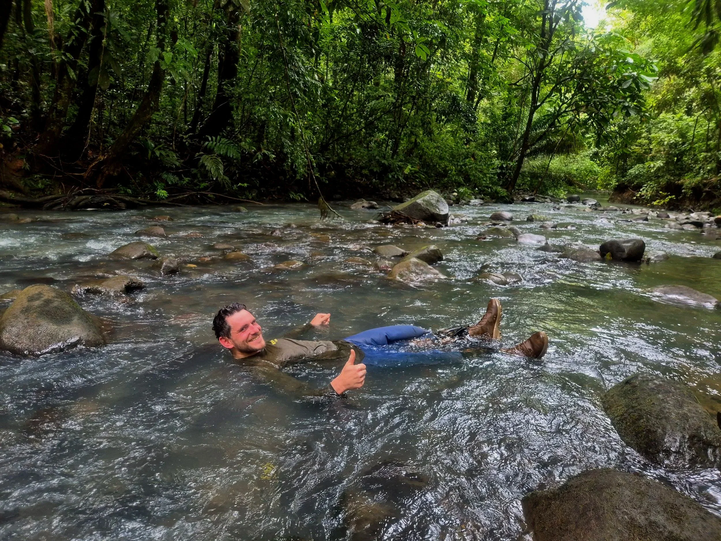 A smiling man in a green jacket and brown boots lying in a shallow river with rocks, giving a thumbs-up, surrounded by lush green trees.