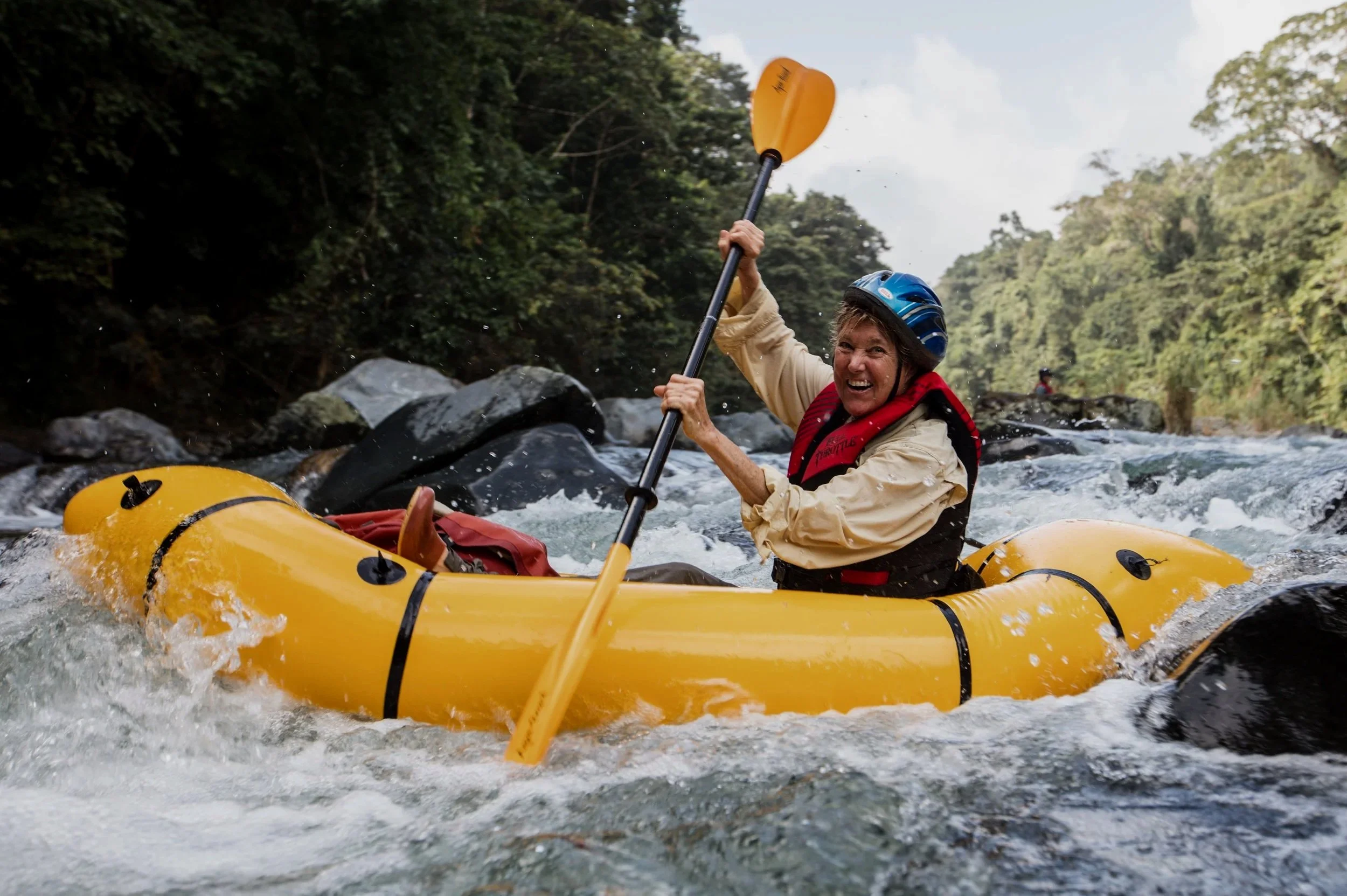 Woman kayaking in a river surrounded by trees, smiling and wearing a blue helmet and red life jacket.