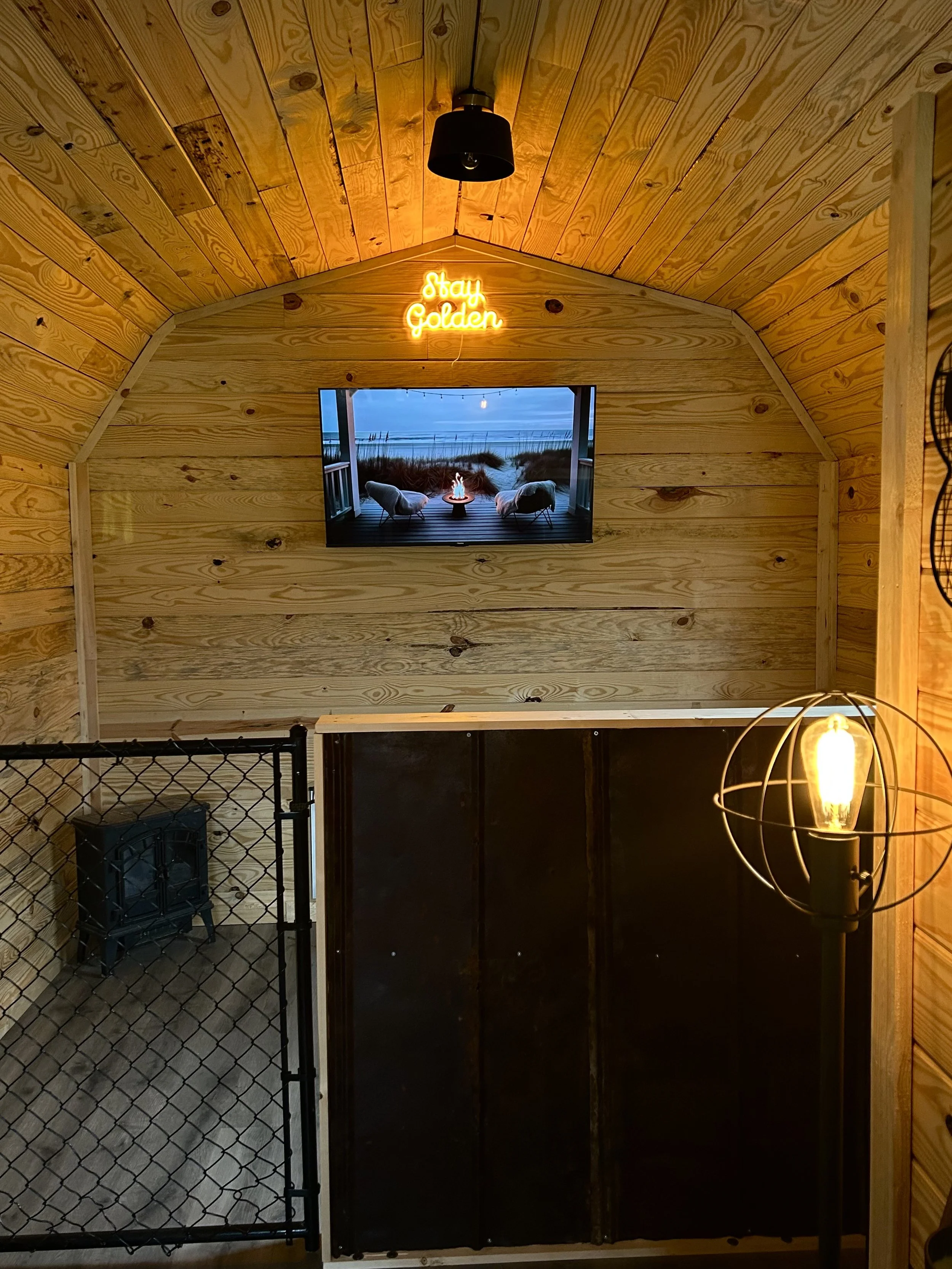 Wood-paneled wall with a mounted TV showing a balcony scene, a neon sign that says 'stay Golden,' a floor lamp with an exposed filament bulb, and a small black stove at the bottom left.