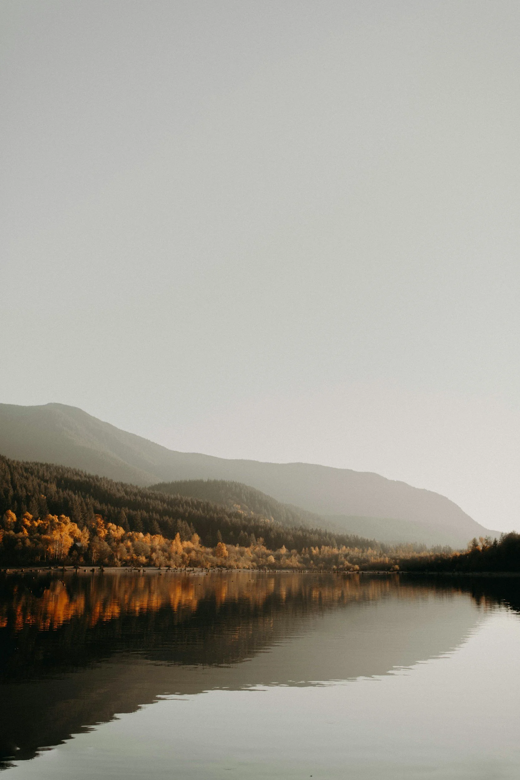 A calm lake with reflections of trees and mountains in the water, during autumn with yellow and orange foliage, under a clear sky.
