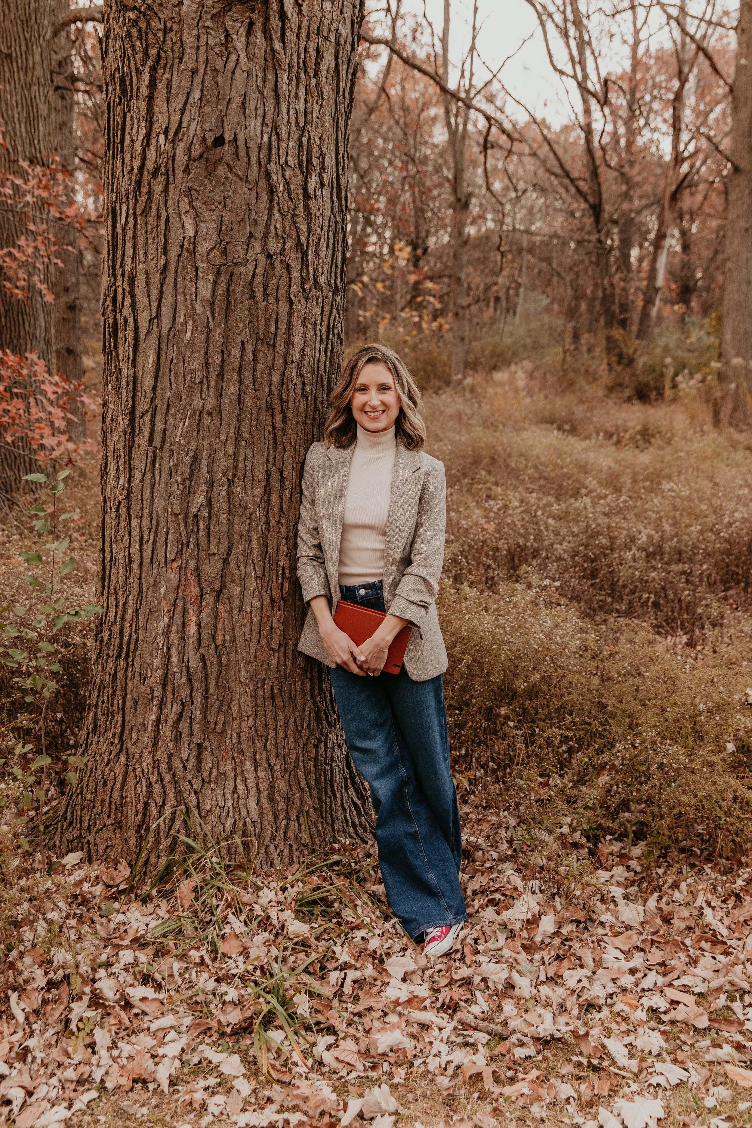A woman standing outdoors in a forest during autumn, smiling, holding a book, and leaning against a large tree trunk, with fallen leaves and trees with autumn foliage in the background.