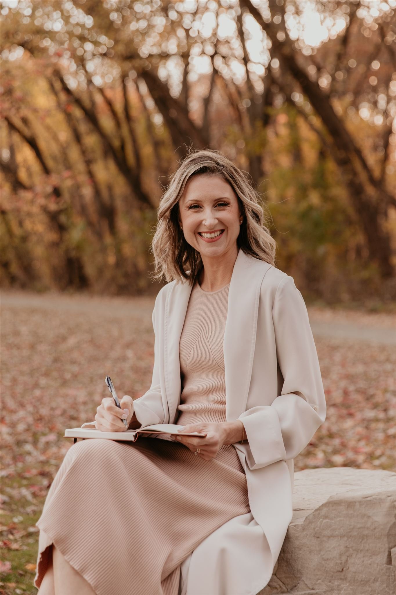 Woman sitting outdoors in autumn, smiling looking at the camera while writing in a notebook.