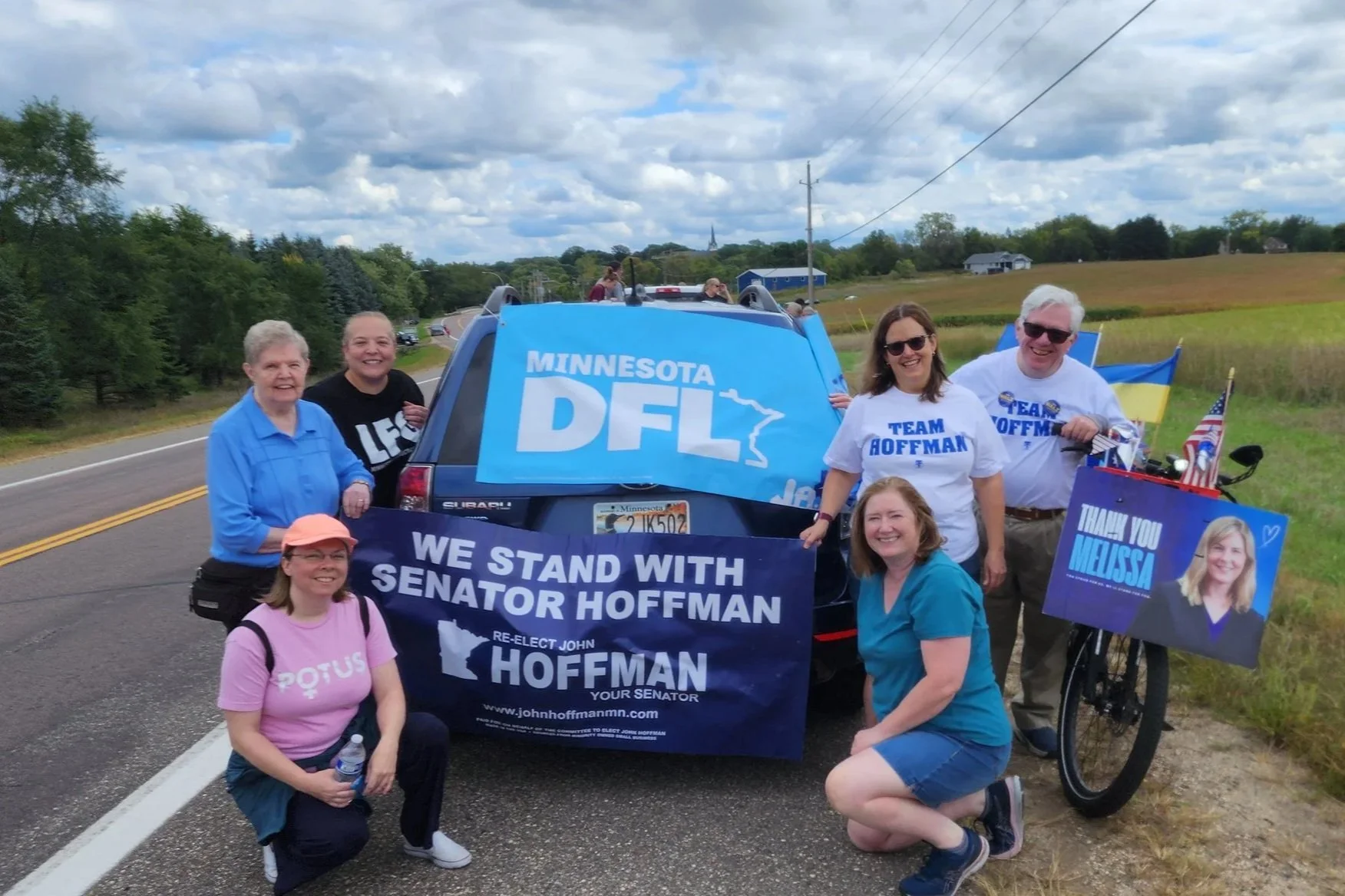 A smiling group sporting banners for Melissa Hortman, John Hoffman, and the Minnesota DFL