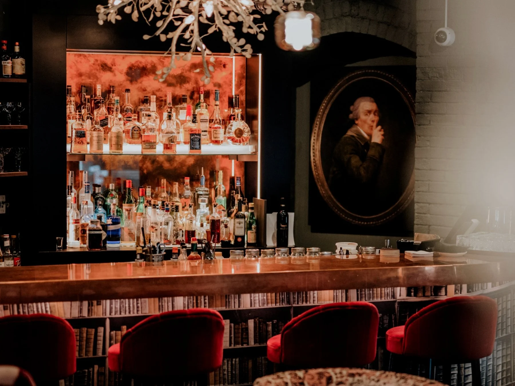 A bar with a well-stocked liquor shelf, red velvet chairs, and a black and white portrait of a person with a finger to their lips, suggesting secrecy or silence.