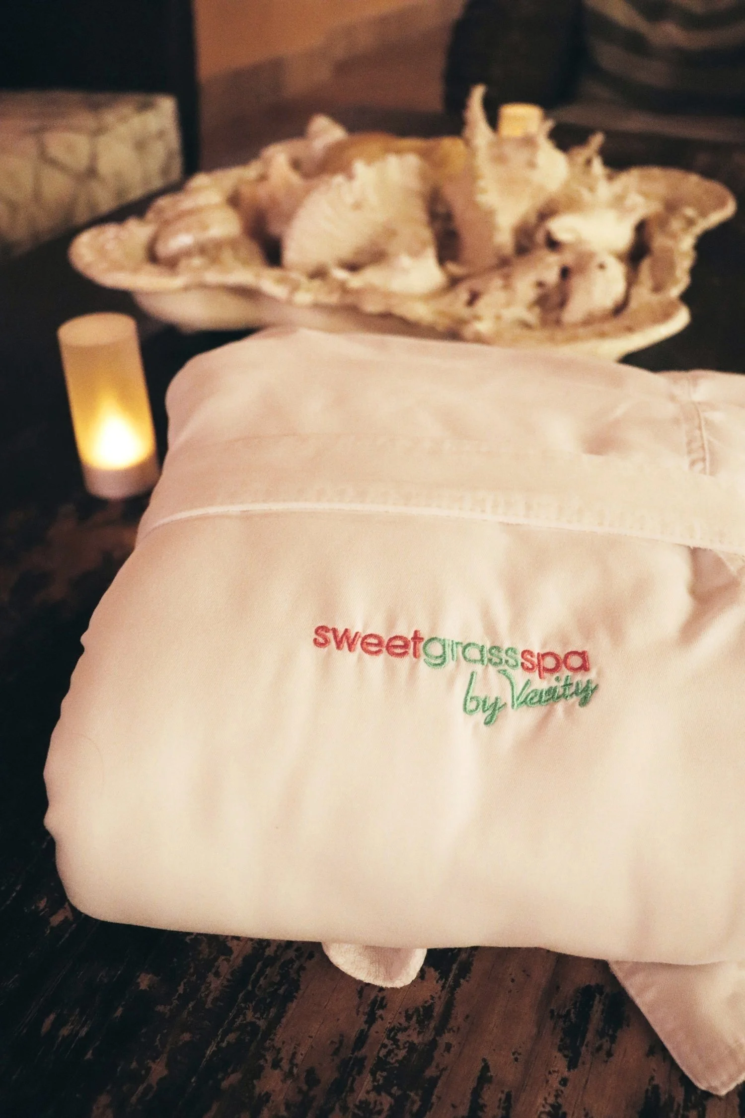 A white towel embroidered with the logo 'sweetgrass spa by vaility' sitting on a wooden surface in a dimly lit spa setting. In the background, there is a large shell bowl filled with rolled towels, and a candle providing soft lighting.