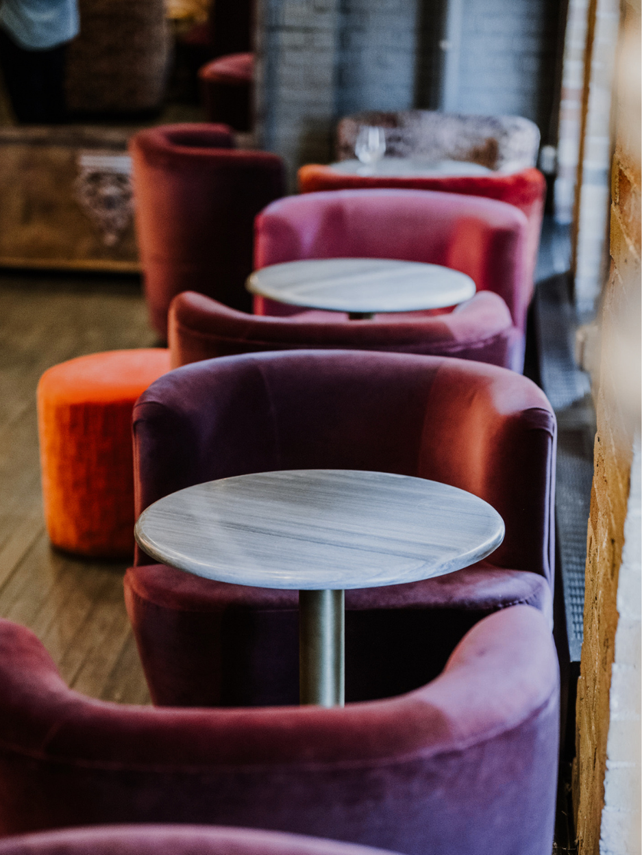 A row of colorful velvet armchairs with round wooden tables in a cozy, dimly lit cafe or lounge.