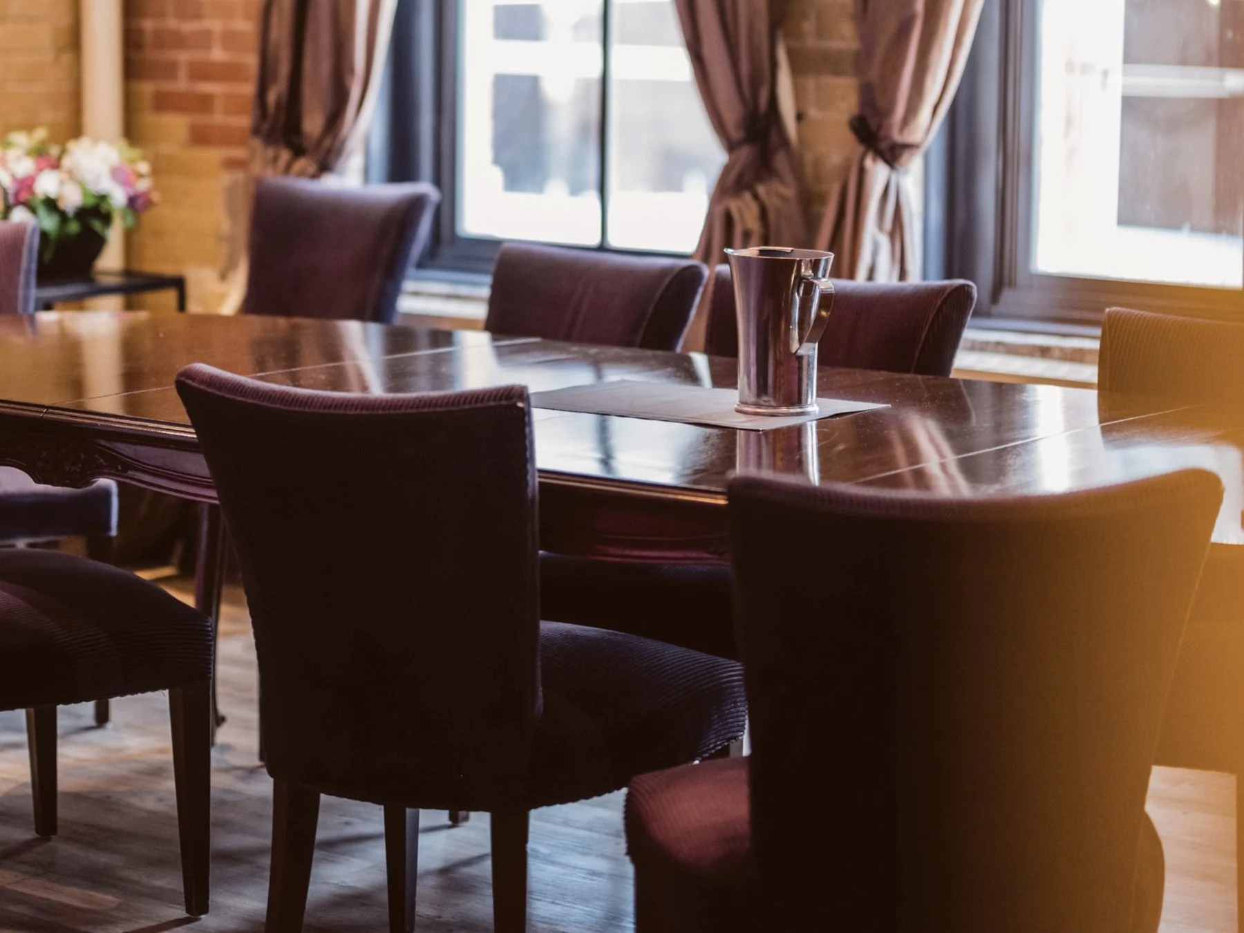 A wooden dining table with six upholstered chairs and a metallic pitcher in the center, set near windows with curtains and a flower arrangement in the background.