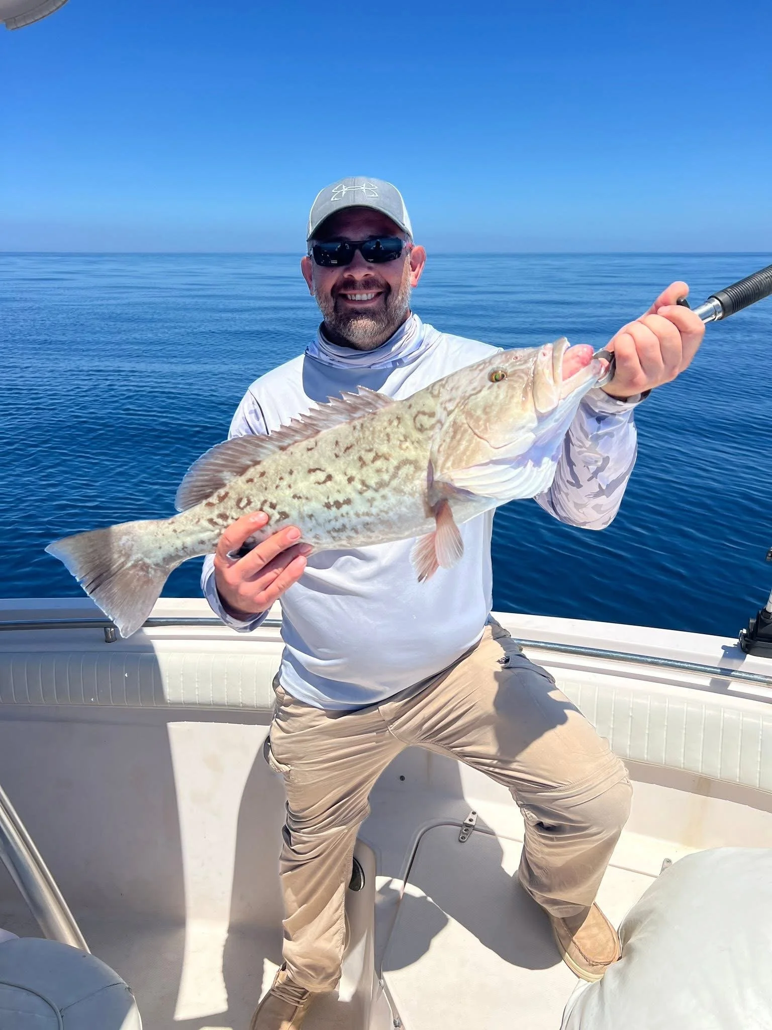 A man wearing sunglasses, a cap, and a long sleeve shirt is sitting on a boat, holding a large fish he caught. The ocean and a clear blue sky are visible in the background.