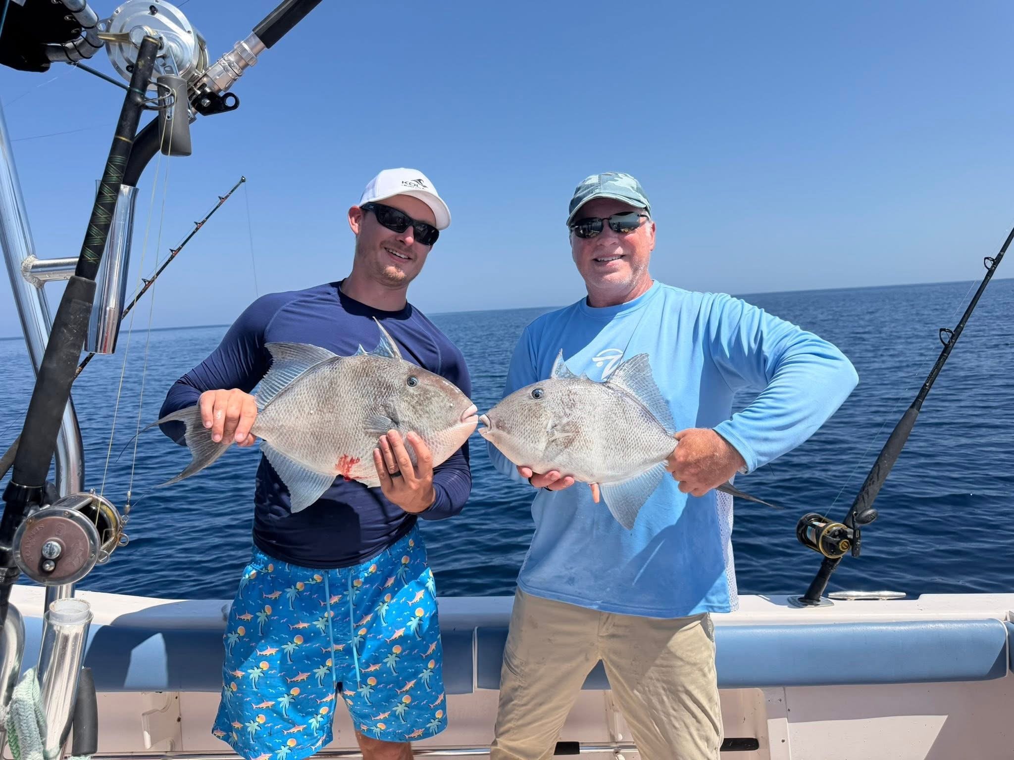 Two men on a boat holding up large fish they caught, with fishing gear and the ocean in the background.