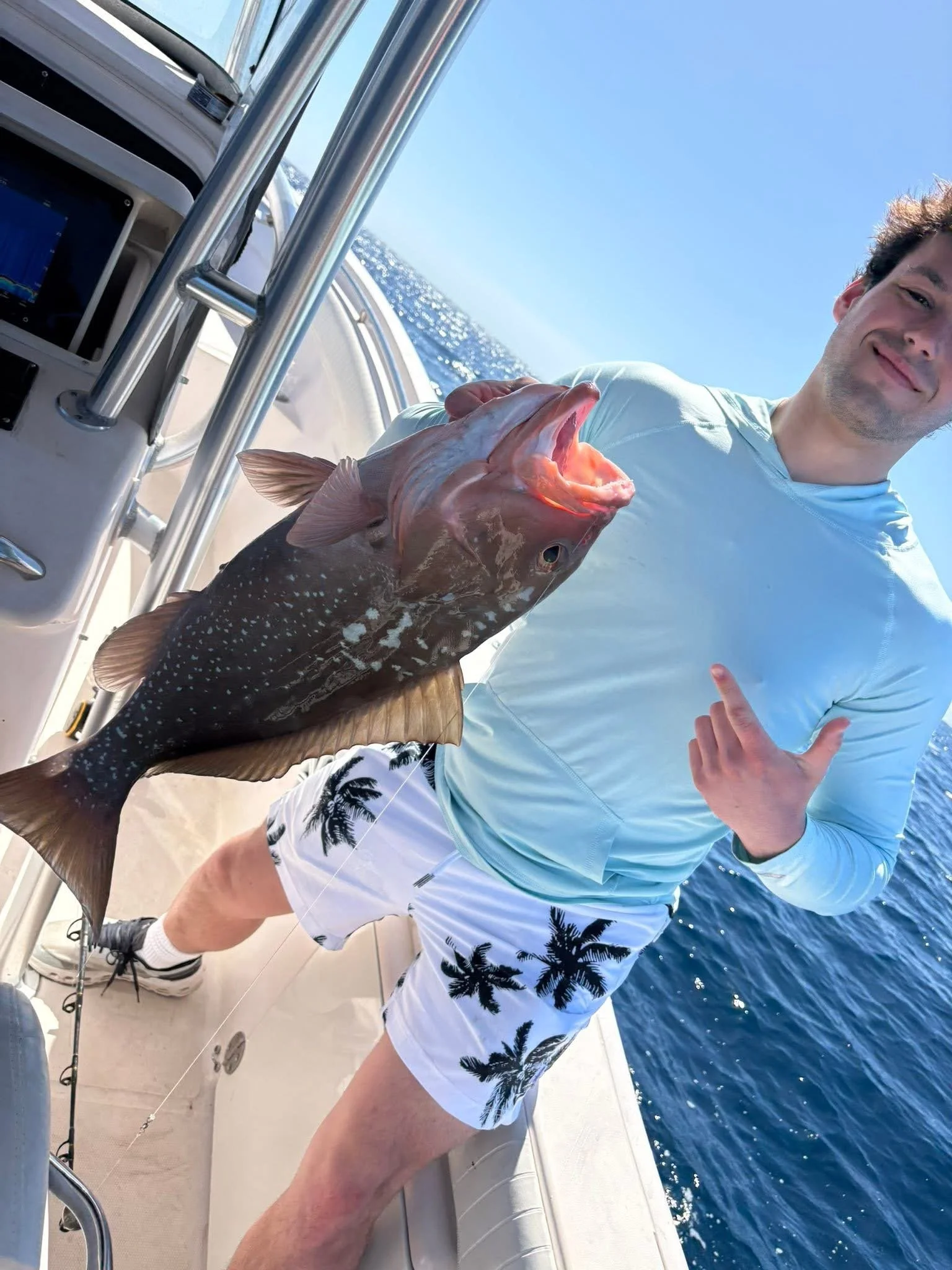 A man on a boat holding a large fish, with the ocean and clear blue sky in the background, making a gesture with his hand.