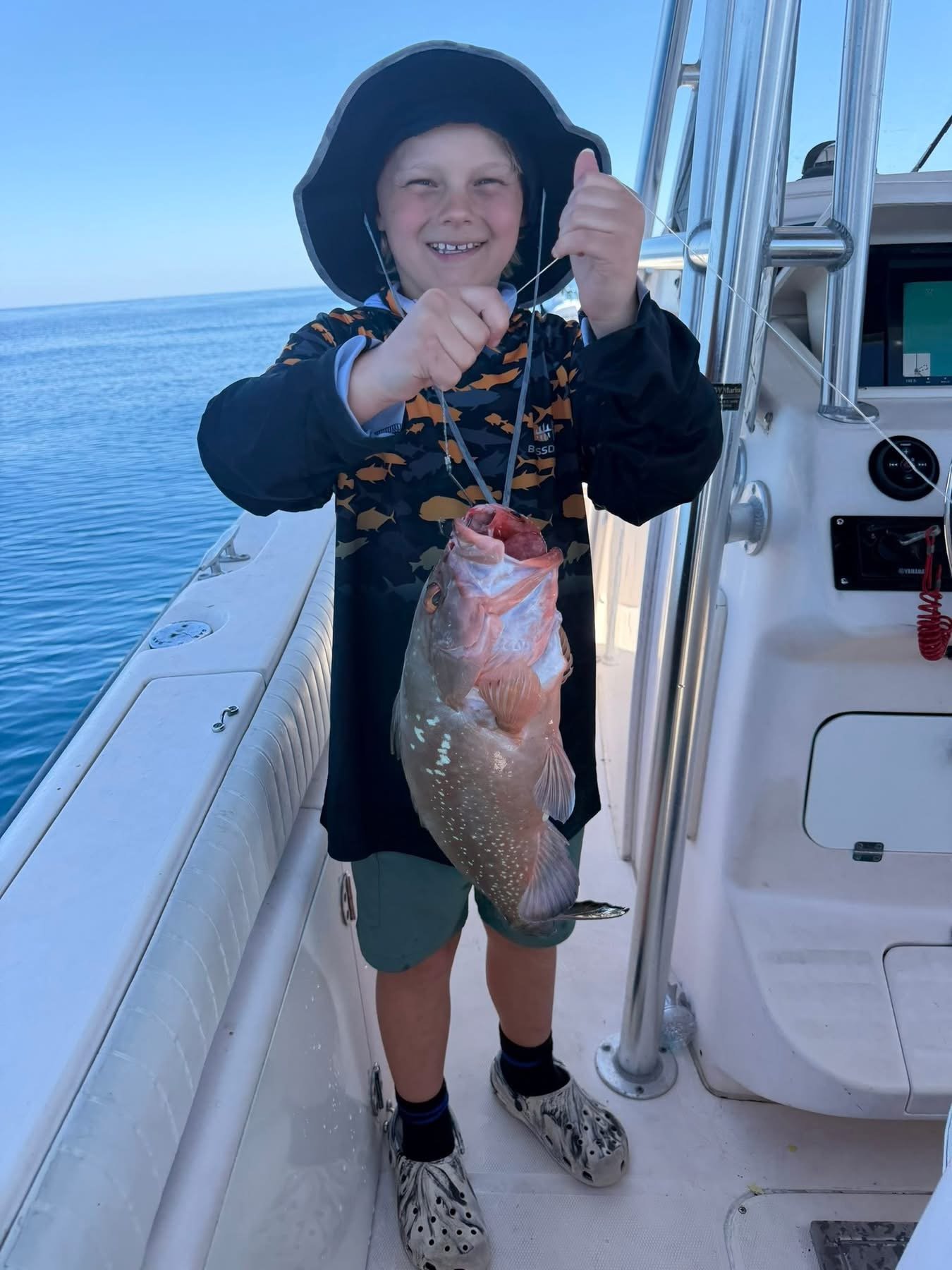 A young boy on a boat holding a large fish, smiling and giving a thumbs up, with the open ocean in the background.