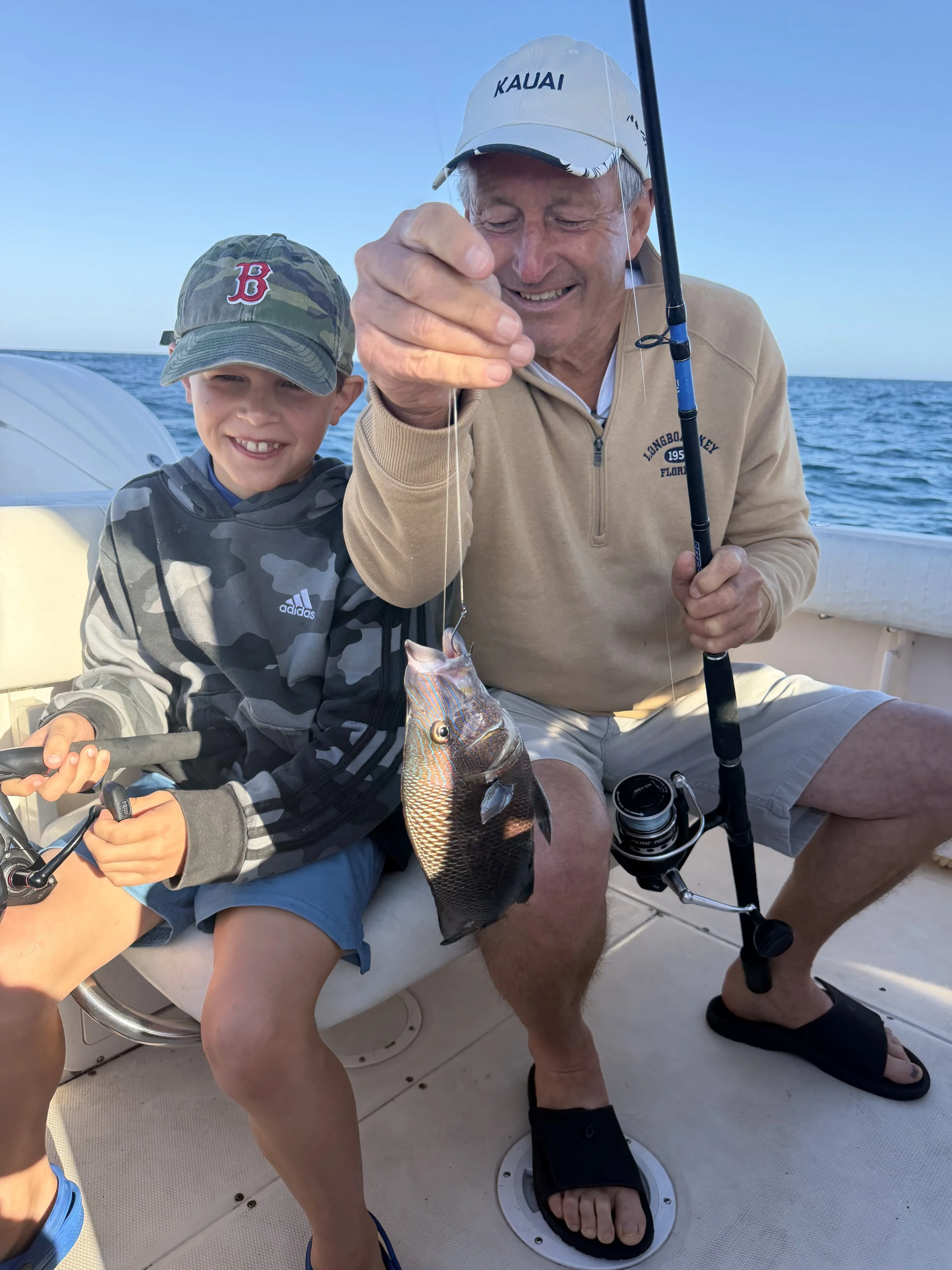 An elderly man and a young boy sitting on a boat, smiling, with the older man holding a fishing rod and a fish they caught, against a background of open water and clear sky.