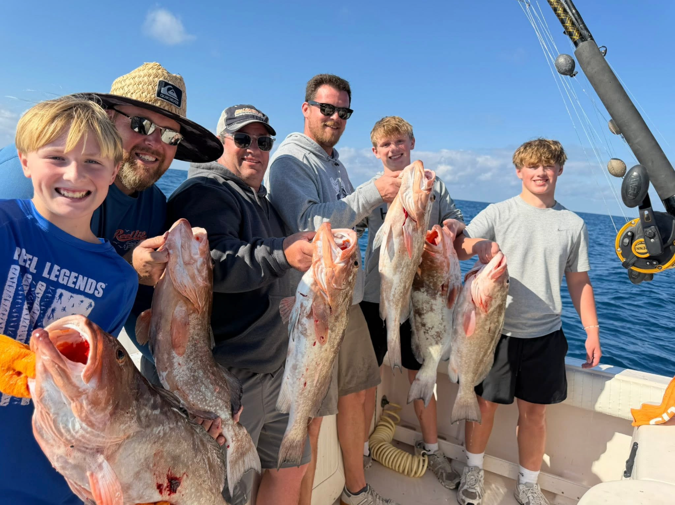 Six person, a family,  on a boat holding large fish they caught during a fishing trip, with a blue ocean and sky in the background.