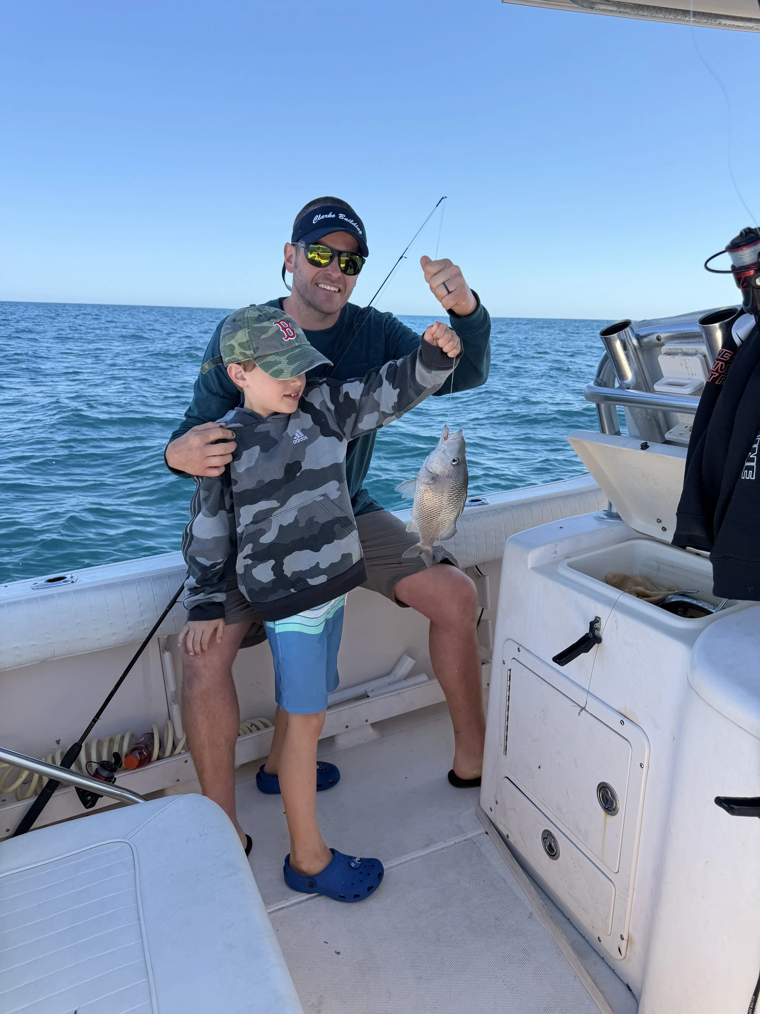 A man and a boy on a boat holding a fish they caught. The man is smiling and wearing sunglasses, a visor, and a dark long sleeve shirt. The boy is wearing a camouflage jacket and a camo cap. The boat is on the water with a clear blue sky overhead.