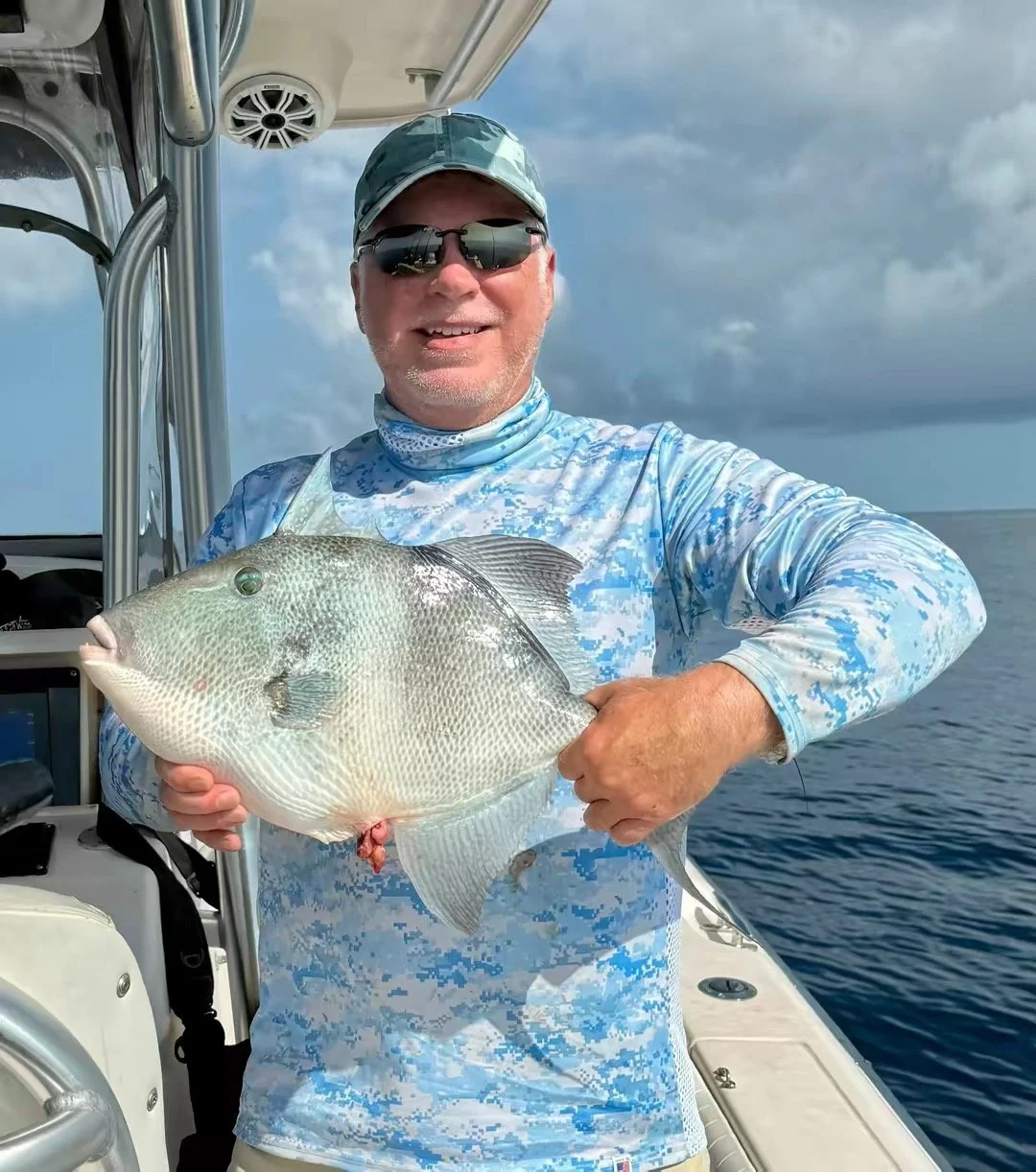 A man wearing sunglasses, a camouflage baseball cap, and a blue camouflage shirt, smiling and holding a large fish onboard a boat at sea under cloudy sky.
