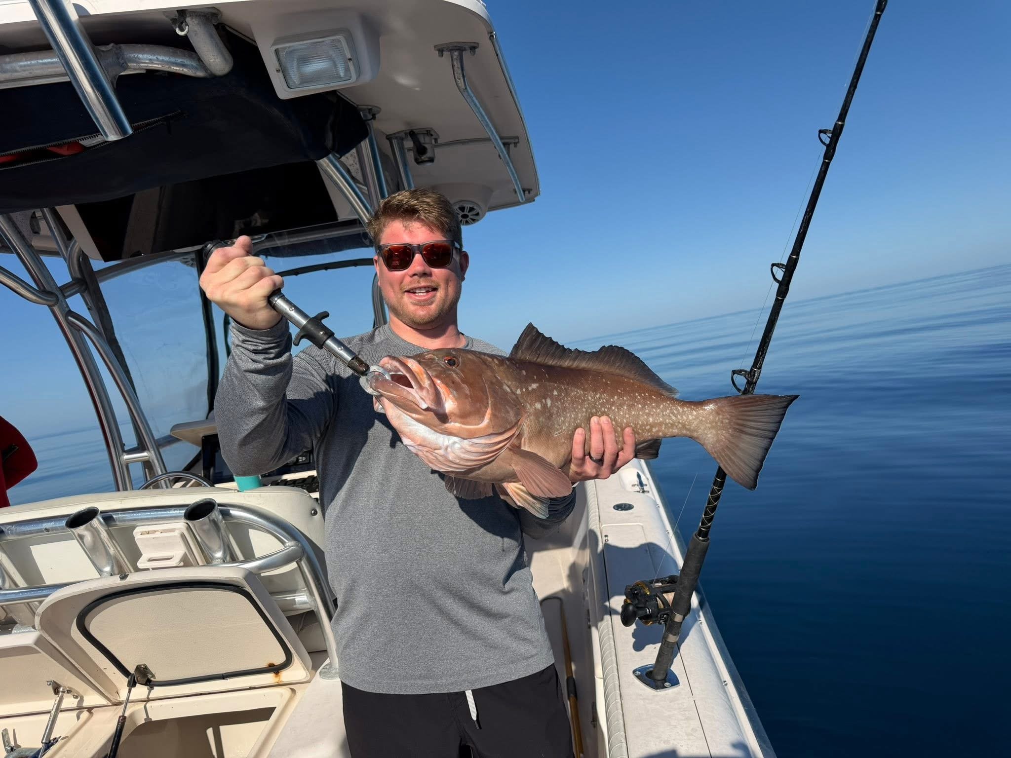 A man wearing sunglasses and a gray long-sleeved shirt on a boat, holding a large fish he caught, with fishing gear and a calm ocean background.