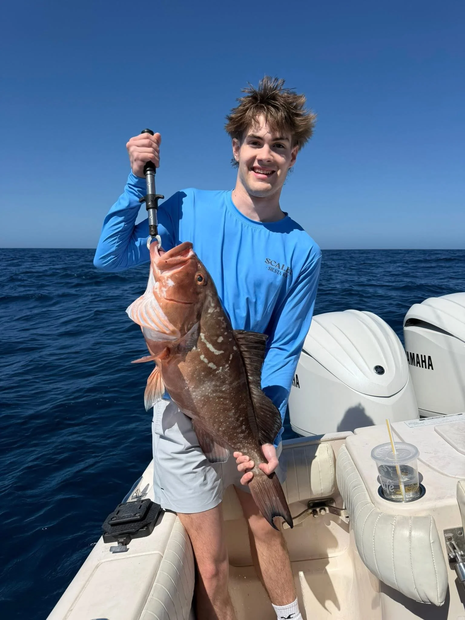A young man with messy hair on a boat, holding a large fish in one hand, with a blue sky and open ocean in the background.