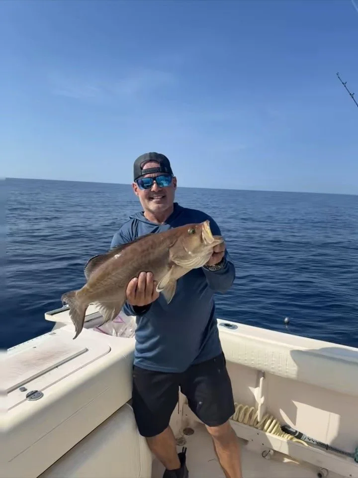 A man on a boat holding a large fish, wearing sunglasses, a baseball cap backwards, a long-sleeve shirt, and shorts, with the ocean and clear sky in the background.