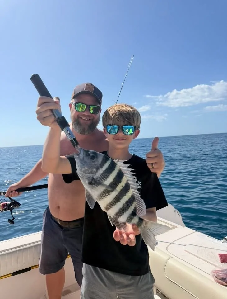 A man and a boy on a boat, smiling and holding a striped fish they caught, with the man giving a thumbs-up and the boy holding fishing gear, ocean in the background on a sunny day.