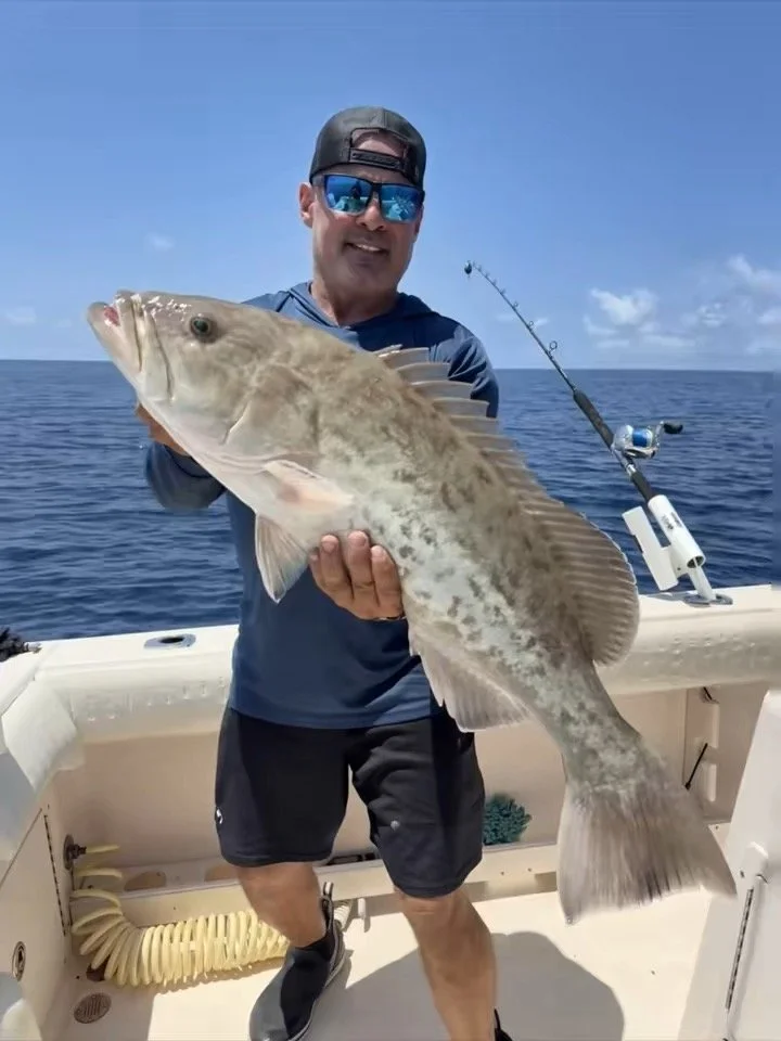 A man wearing a baseball cap, sunglasses, a blue jacket, and shorts is smiling and holding a large fish on a boat out at sea. The background shows the ocean and a blue sky with some clouds.
