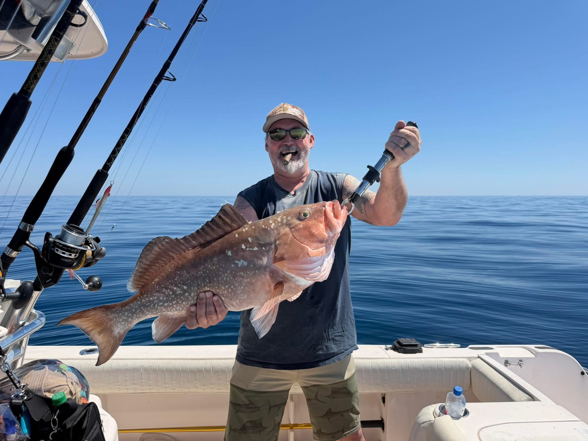 A man on a boat holding a large fish he has caught, with fishing rods visible on the boat and a calm ocean in the background under a blue sky.
