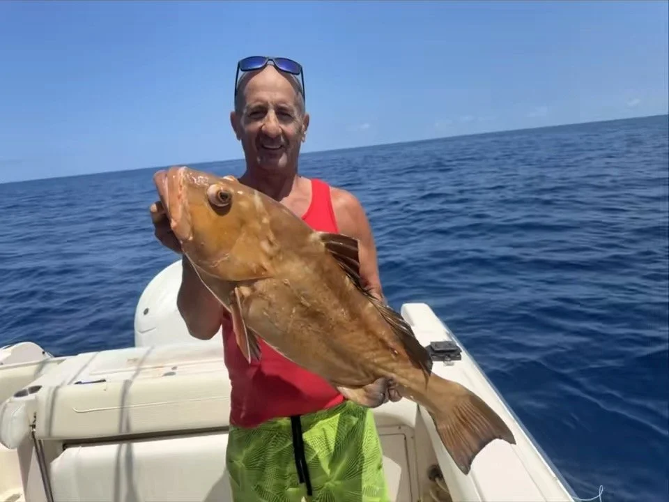 A man on a boat holding a large fish in the ocean with a blue sky in the background.