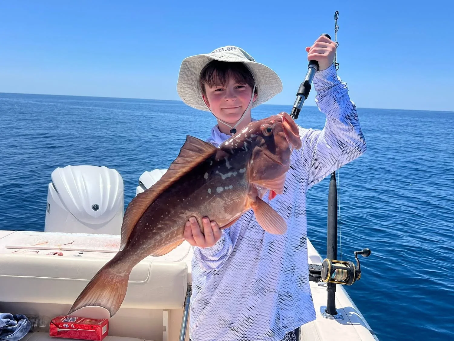 Boy on a boat holding a large fish he caught while fishing at sea. He is wearing a wide-brimmed hat and a long sleeve shirt, with fishing gear in the background.