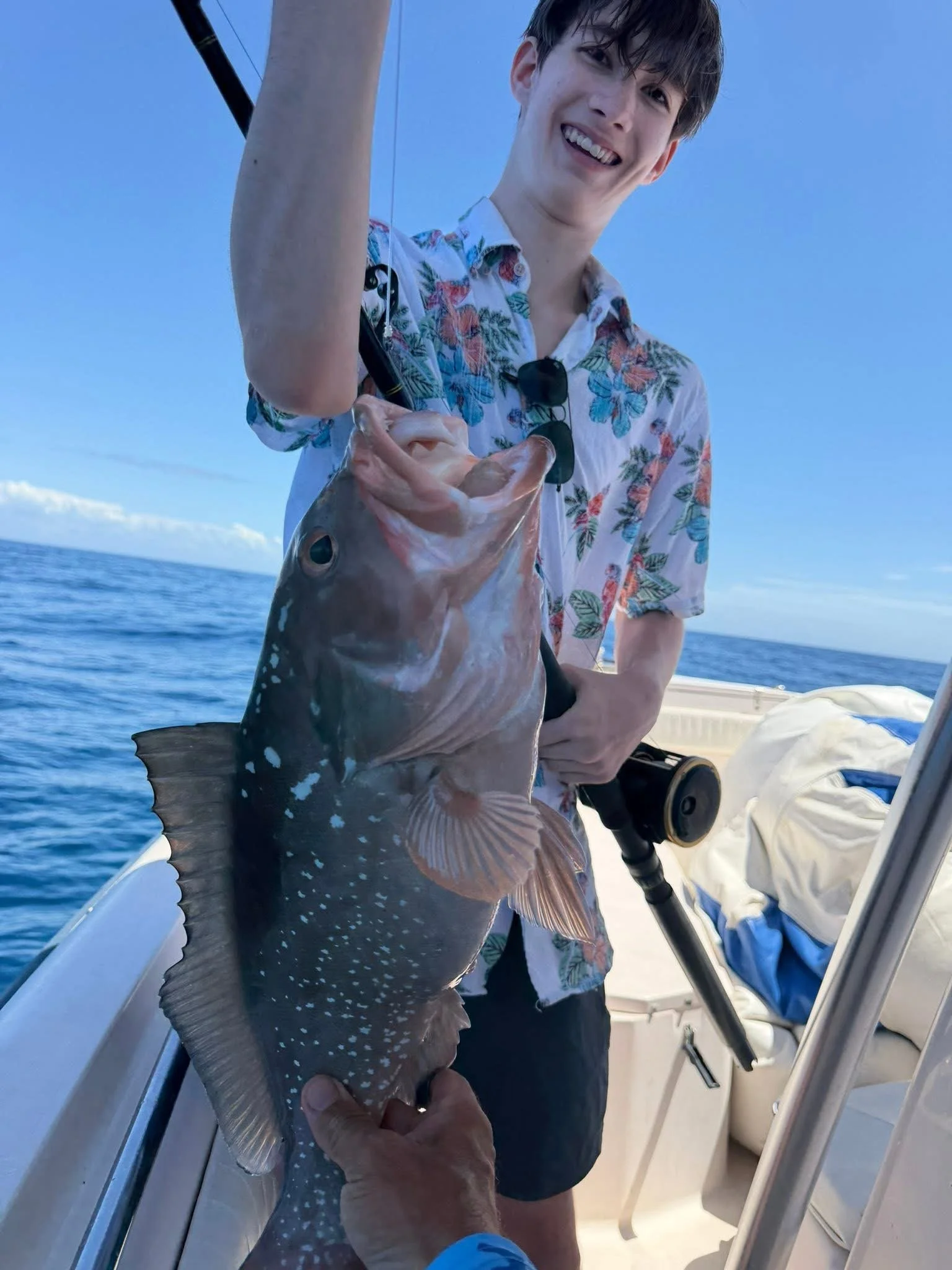 A smiling young man in a tropical shirt holding a fish on a boat, with the ocean and blue sky in the background.